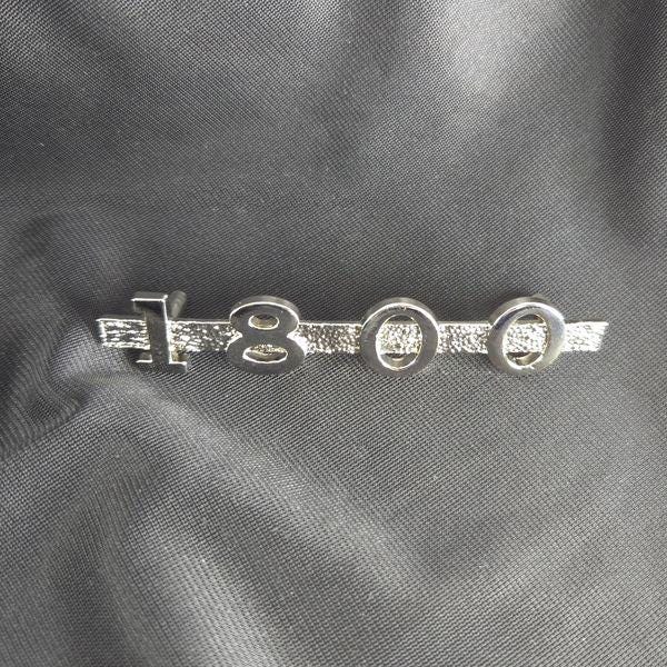 Shiny metallic MGA 1800 emblem on black cloth background, angled to emphasize reflective electroformed nickel plating and clean edges.