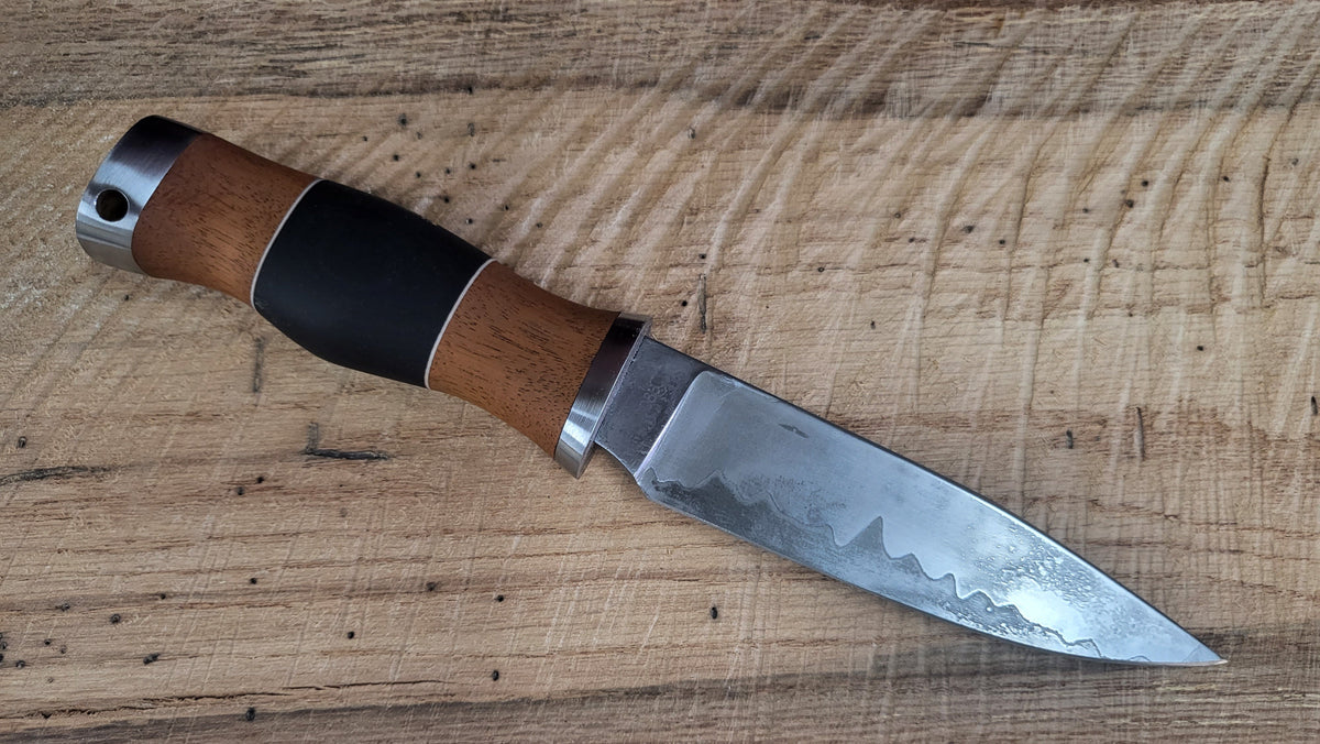 "Finished hidden‑tang knife in Go Mai forge weld from 1520, 1095, and railroad spikes with visible hamon, mahogany handle with blue micarta center and steel pommel, displayed on wooden background as completed custom commission piece