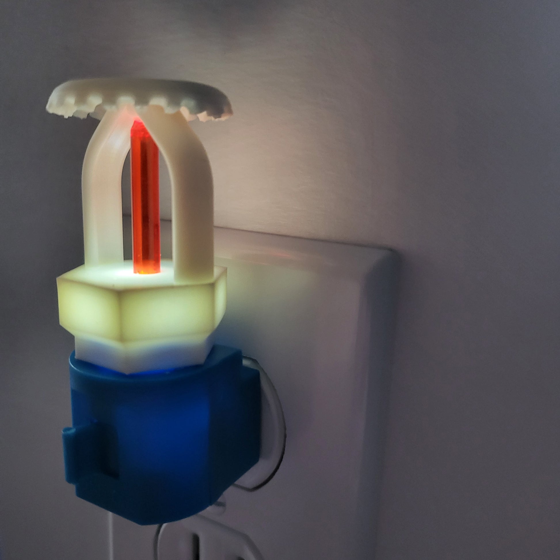 Right‑side view of fire sprinkler night light glowing softly in dim room; white upright head with red bulb sits on blue base with manual switch, bringing a touch of fire protection pride to home décor.