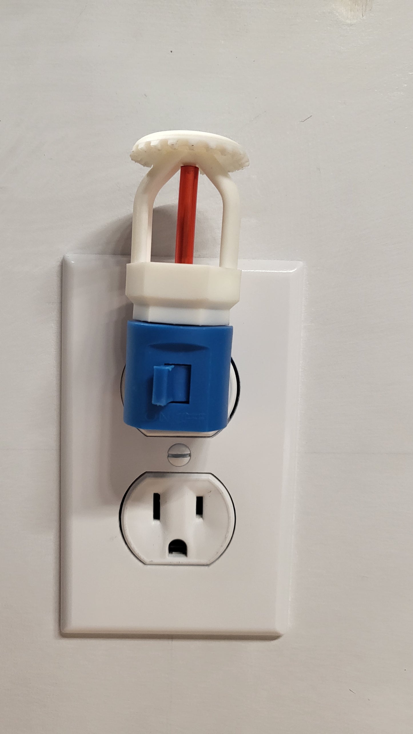 Straight‑on view of white upright fire sprinkler night light with red bulb on blue base with switch, plugged into wall outlet in off position against clean white wall.