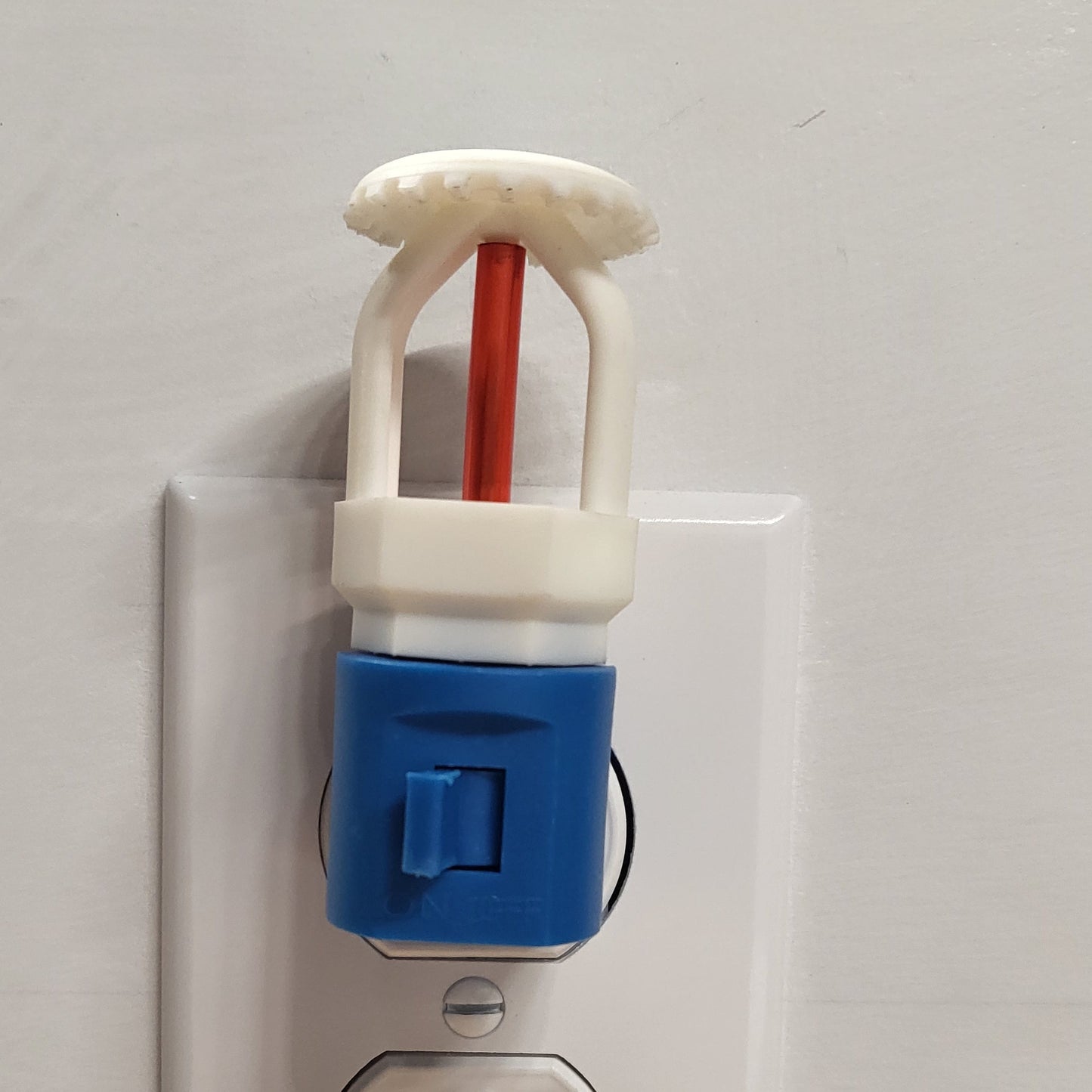 Straight‑on view of white upright fire sprinkler night light with red bulb on blue base with switch, plugged into wall outlet in off position against clean white wall.