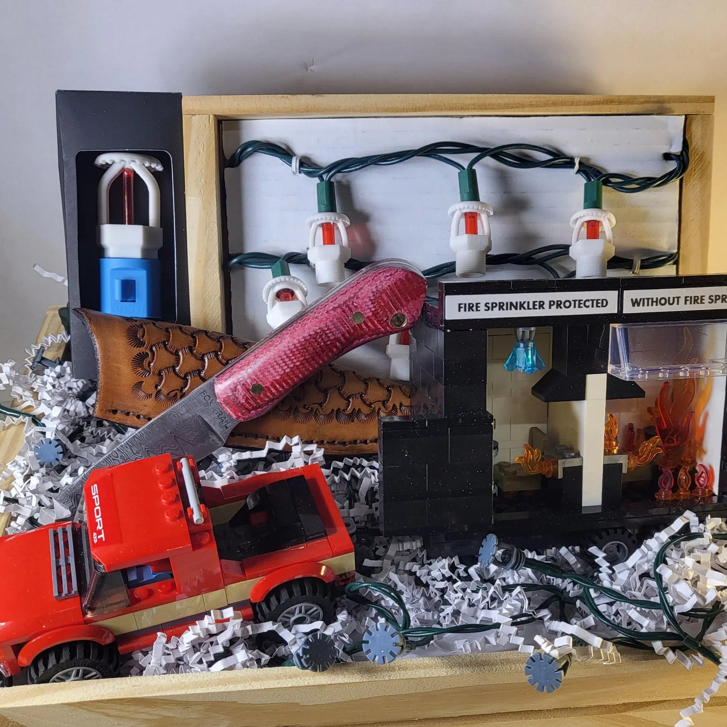 Gift basket created for FFSA’s Buddy Dewar Tournament raffle featuring Fox Trail Craftworks fire protection products — white string lights, building block burn trailer with truck, upright sprinkler night light, and a hand‑forged knife with fire hose micarta‑style handle.