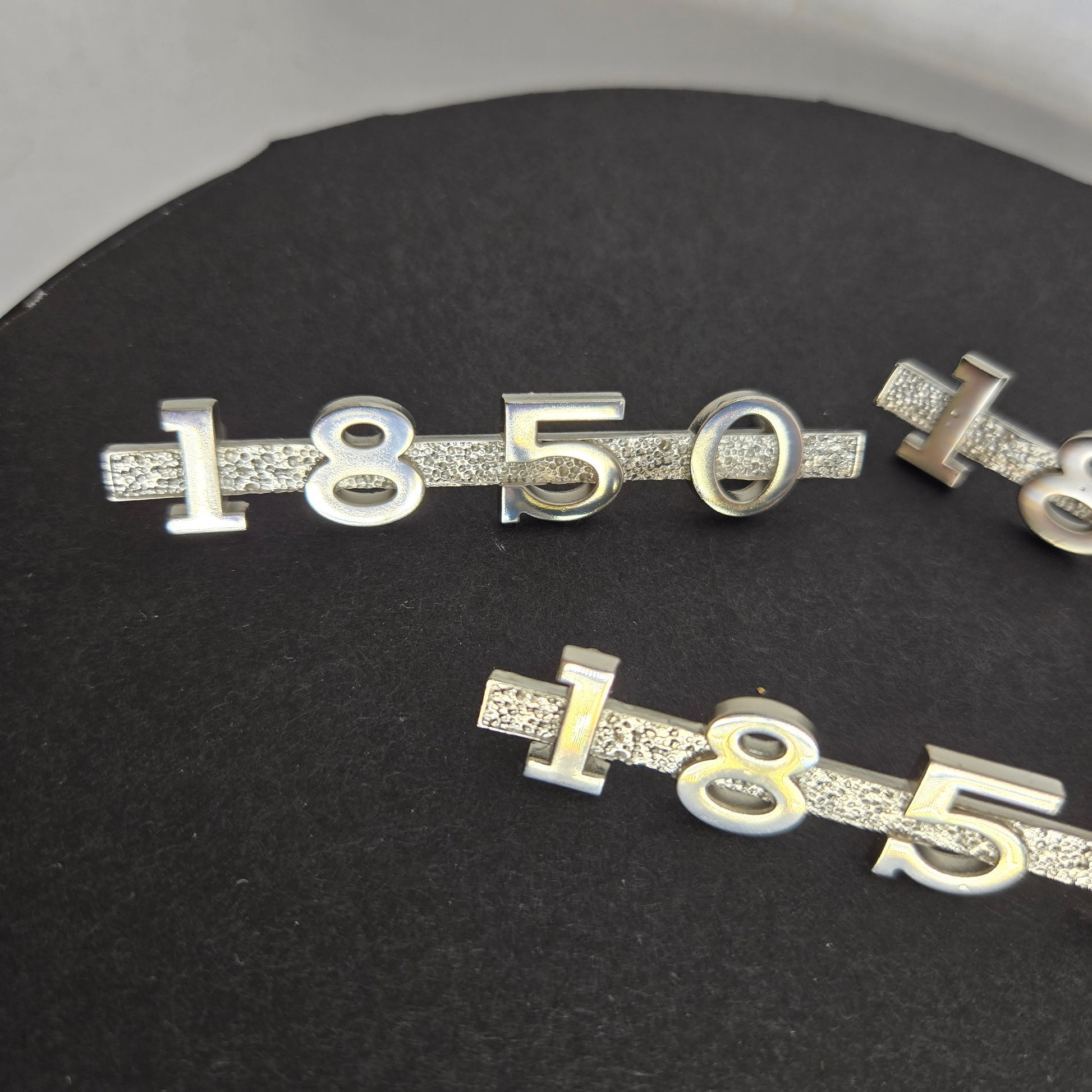 Alternate angle of three MGA 1850 emblems with mounting pins on black disc and white background, emphasizing reflective finish and craftsmanship.