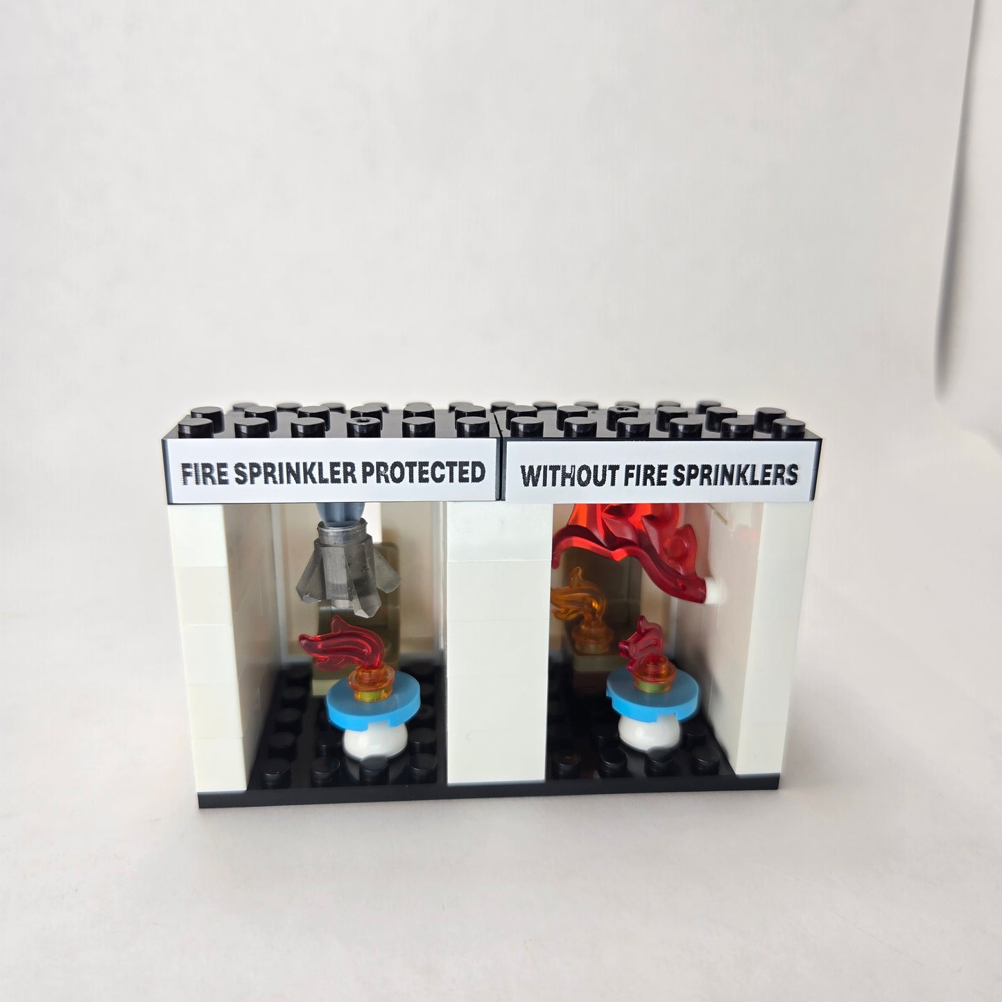 Mini fire sprinkler demo kit showing two side‑by‑side rooms — one labeled ‘Fire Sprinkler Protected’ with an activated sprinkler featuring a smoky crystal to humorously represent real sprinkler water, and the other labeled ‘Without Fire Sprinklers’ engulfed in flames to illustrate flashover.
