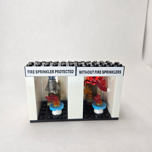 Mini fire sprinkler demo kit showing two side‑by‑side rooms — one labeled ‘Fire Sprinkler Protected’ with an activated sprinkler featuring a smoky crystal to humorously represent real sprinkler water, and the other labeled ‘Without Fire Sprinklers’ engulfed in flames to illustrate flashover.