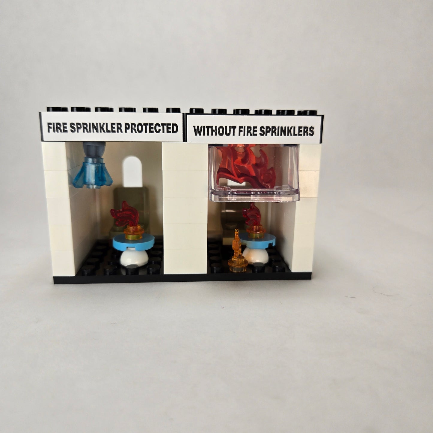 Mini fire sprinkler demo kit built from blocks, showing two side‑by‑side rooms with chair and table — one labeled ‘Fire Sprinkler Protected’ with a sprinkler element and blue crystal water drop, and the other labeled ‘Without Fire Sprinklers’ enclosed in flames to illustrate flashover.