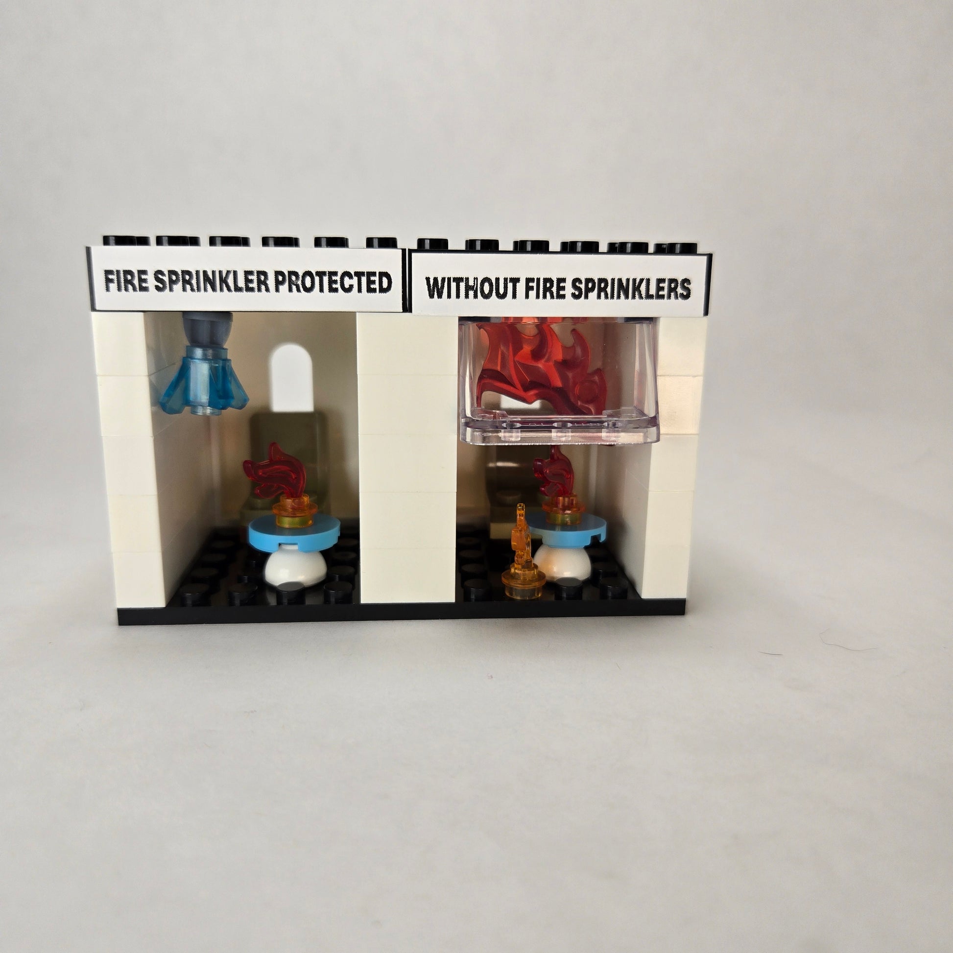 Mini fire sprinkler demo kit built from blocks, showing two side‑by‑side rooms with chair and table — one labeled ‘Fire Sprinkler Protected’ with a sprinkler element and blue crystal water drop, and the other labeled ‘Without Fire Sprinklers’ enclosed in flames to illustrate flashover.
