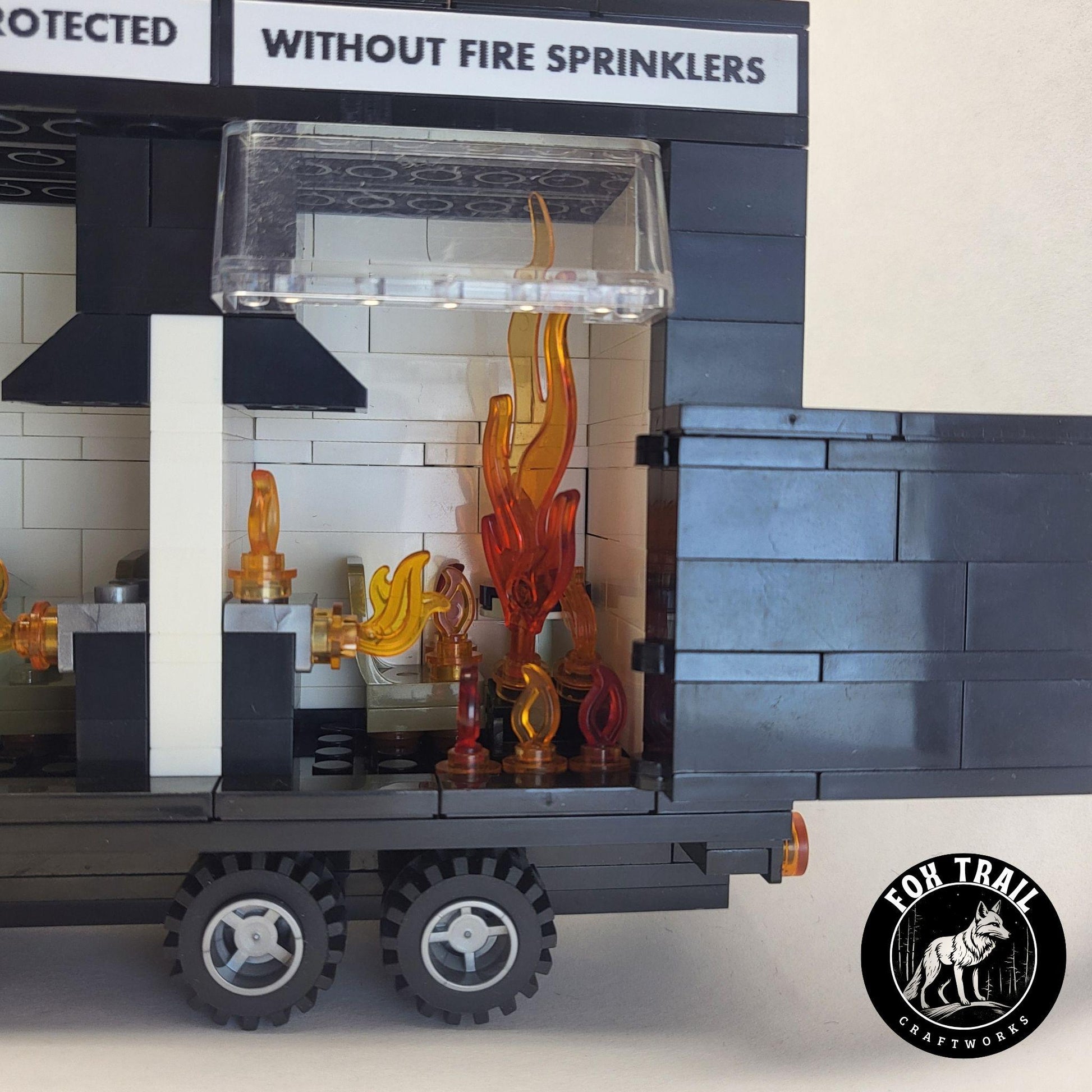 Detailed view of the ‘Without Fire Sprinklers’ side of the Lego‑compatible burn trailer, highlighting rolling wheels, windscreen, and multiple flame elements illustrating flashover inside the small kitchen scene.