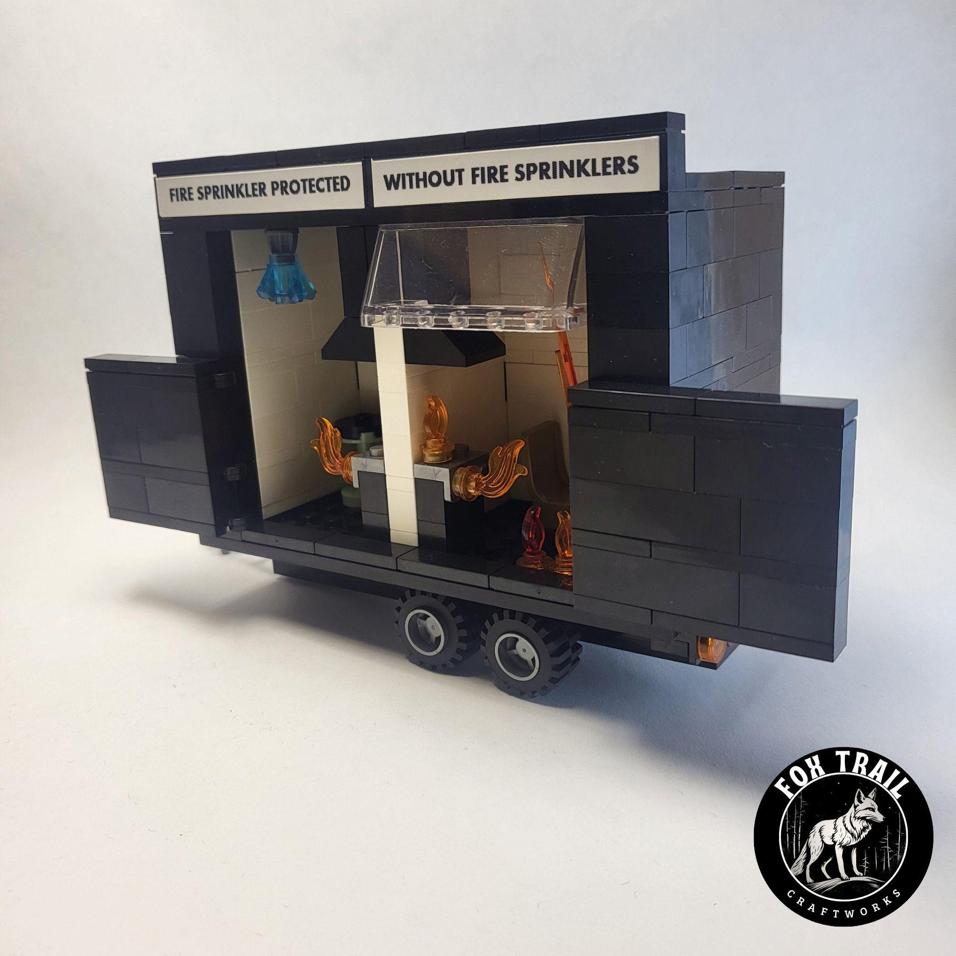 Right‑side view of Lego‑compatible burn trailer with doors open, revealing identical kitchen setups labeled for fire sprinkler protection versus no sprinklers, complete with stove, table, and blue crystal sprinkler activation detail.
