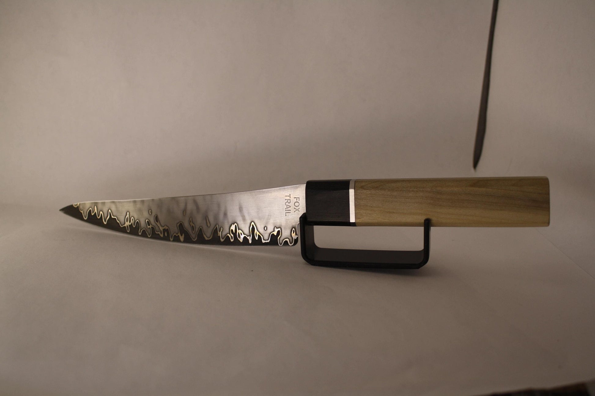 Opposite side of San‑Mai kitchen knife on white background with black stand, displaying symmetrical flame‑like pattern and curved spine for ergonomic control.