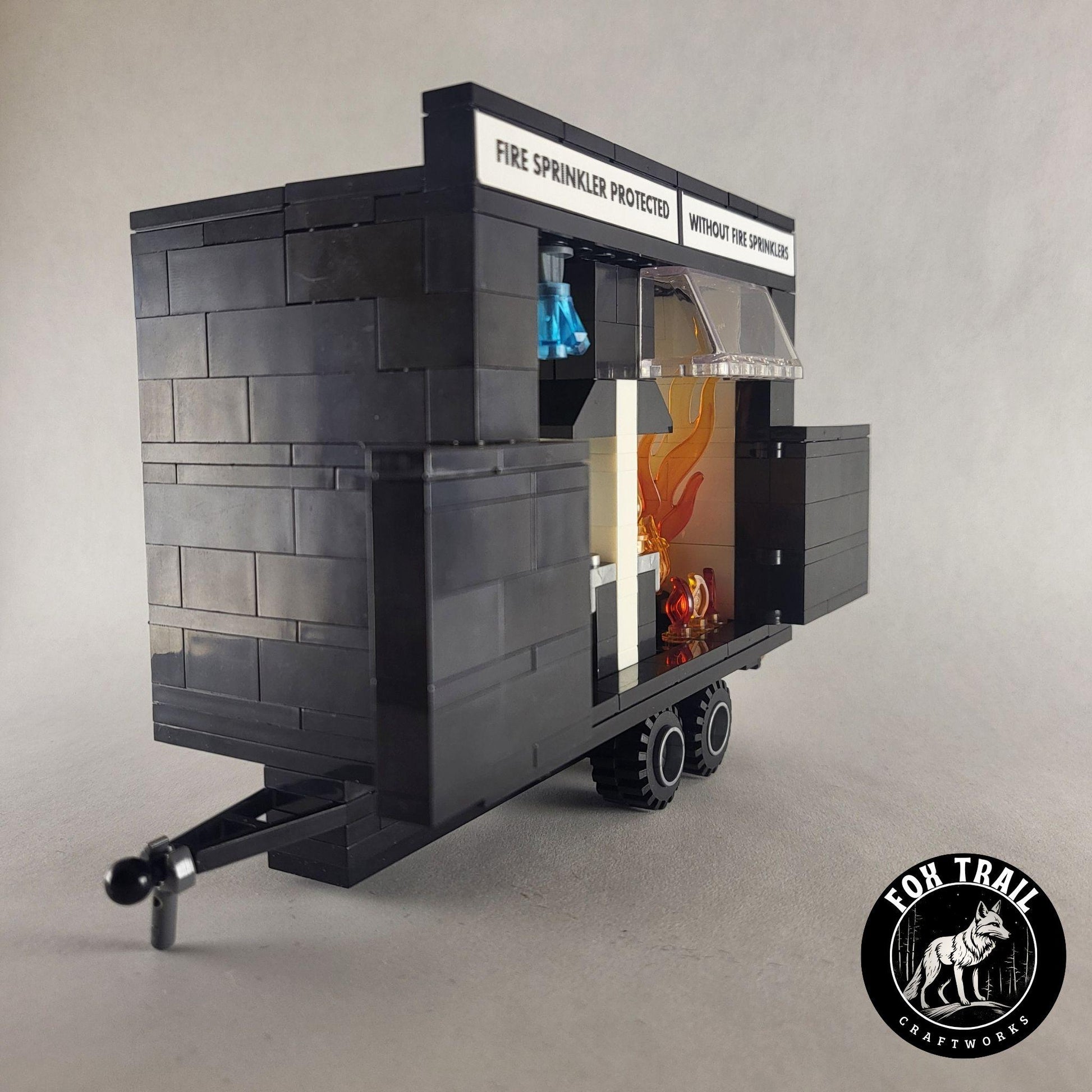 Left‑side view of educational burn trailer built from blocks, doors open to show kitchen interiors labeled for sprinkler protection versus no sprinklers, with realistic details like coupler, wheels, windscreen, and activated blue crystal sprinkler.
