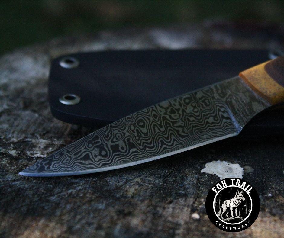 Close‑up of Damascus blade edge from Fox Trail Craftworks neck knife on log with grass background; sharp cutting edge in focus with black Kydex sheath visible behind.