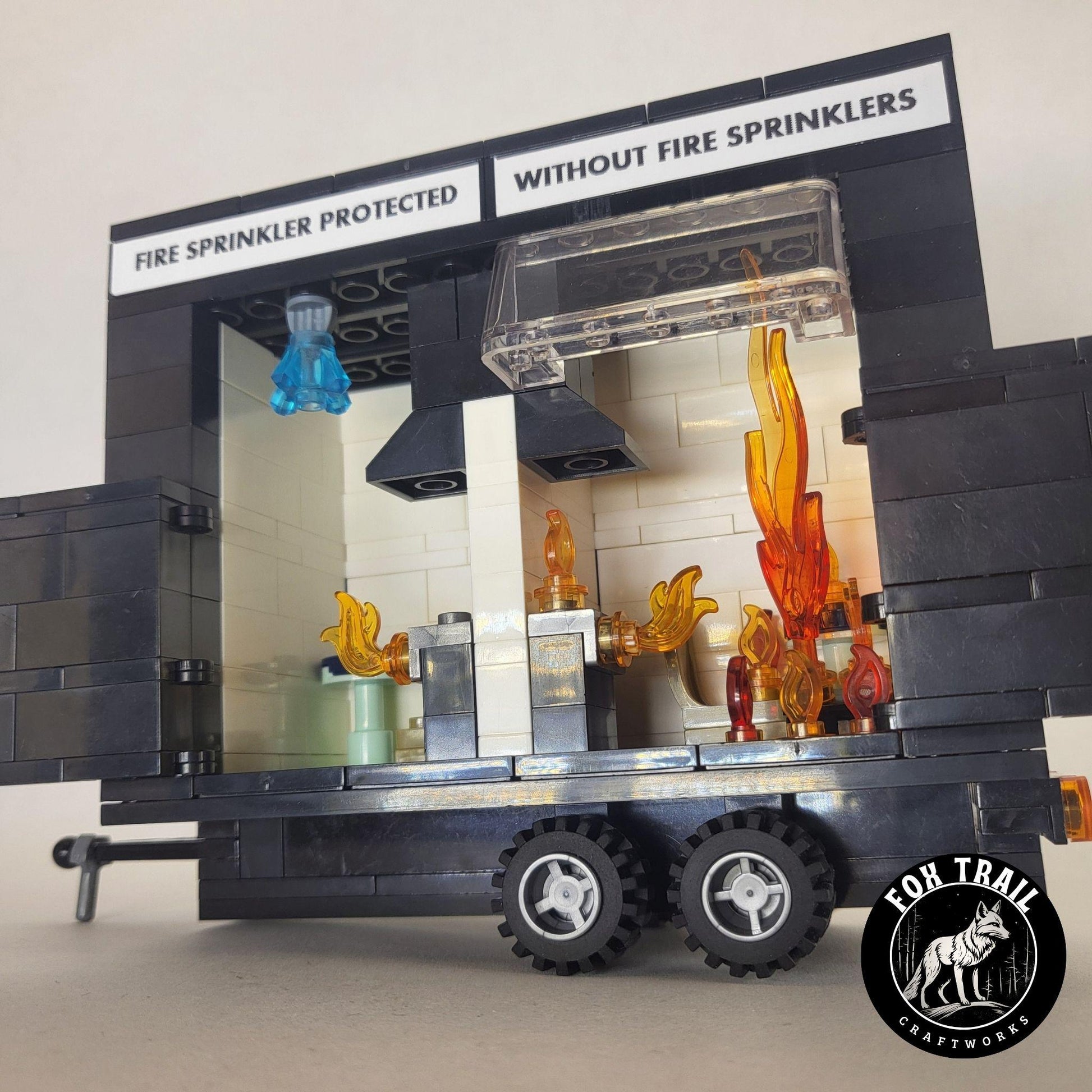 Complete building block burn trailer display compatible with Lego, showing open doors, trailer coupler, tail lights, and labeled sides — ‘Fire Sprinkler Protected’ and ‘Without Fire Sprinklers.’ Kitchen scenes inside feature a working sprinkler with blue water crystal and protective windscreen for realistic fire safety education.