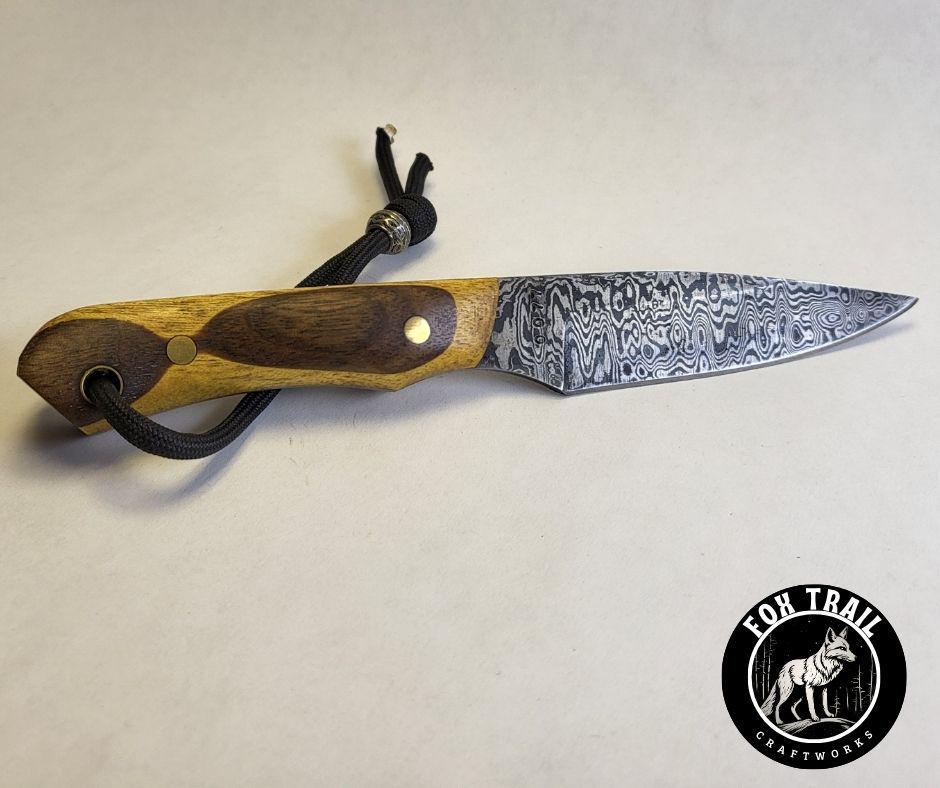 Flat lay of handmade Damascus neck knife on white background, showcasing random Damascus pattern blade, full tang construction, and black wrist thong with bead.