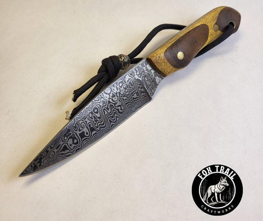 Top‑front angled view of Fox Trail Craftworks Damascus neck knife on white background, showing makers mark near handle, Yellowheart and walnut stacked grip, and black wrist thong with bead.