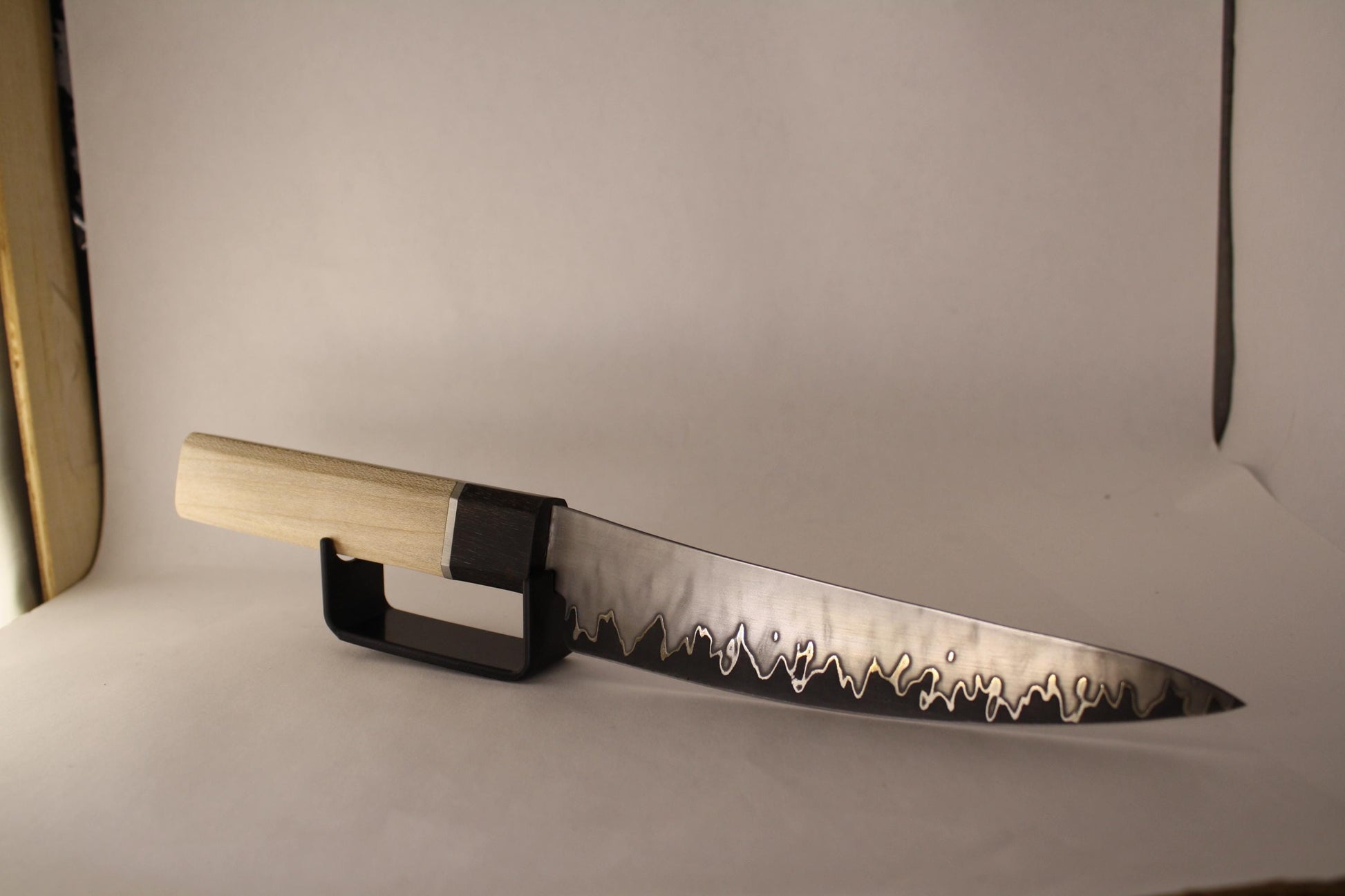 San‑Mai utility knife on white background with black stand, showing gently curved spine and flame‑like blade pattern forged from 1095 core, nickel layers, and stainless jacket.
