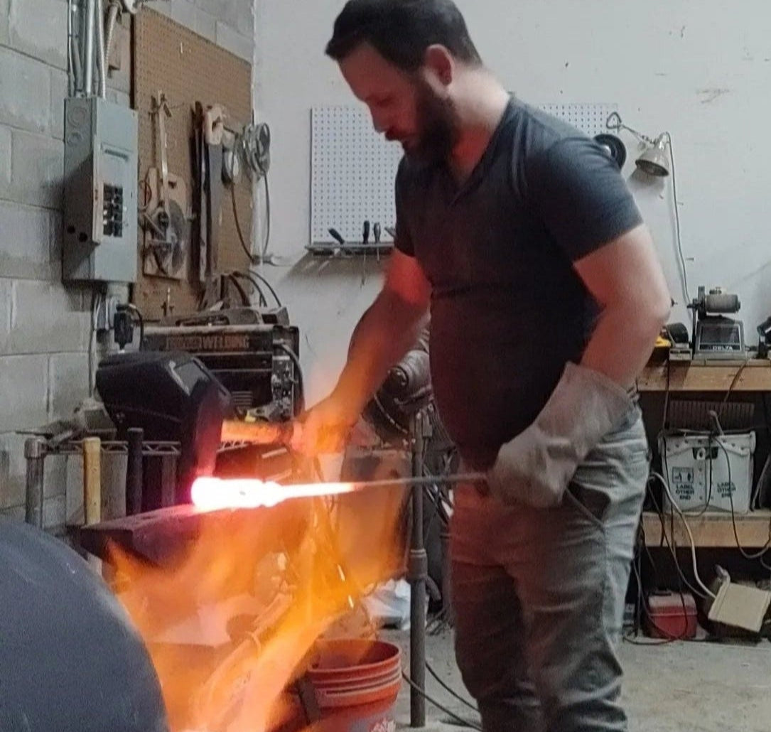 Chris forging a Damascus billet on an anvil in his workshop, flames from the forge rising in the foreground as he hammers the heated steel with a gloved hand.