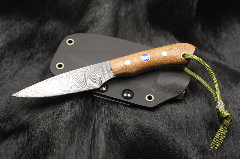 Close‑up of custom Damascus neck knife showing intricate blade pattern and custom blue mosaic logo pin, resting on black Kydex sheath. Knife displayed on black fur background, part of a commissioned set for a local life safety company.