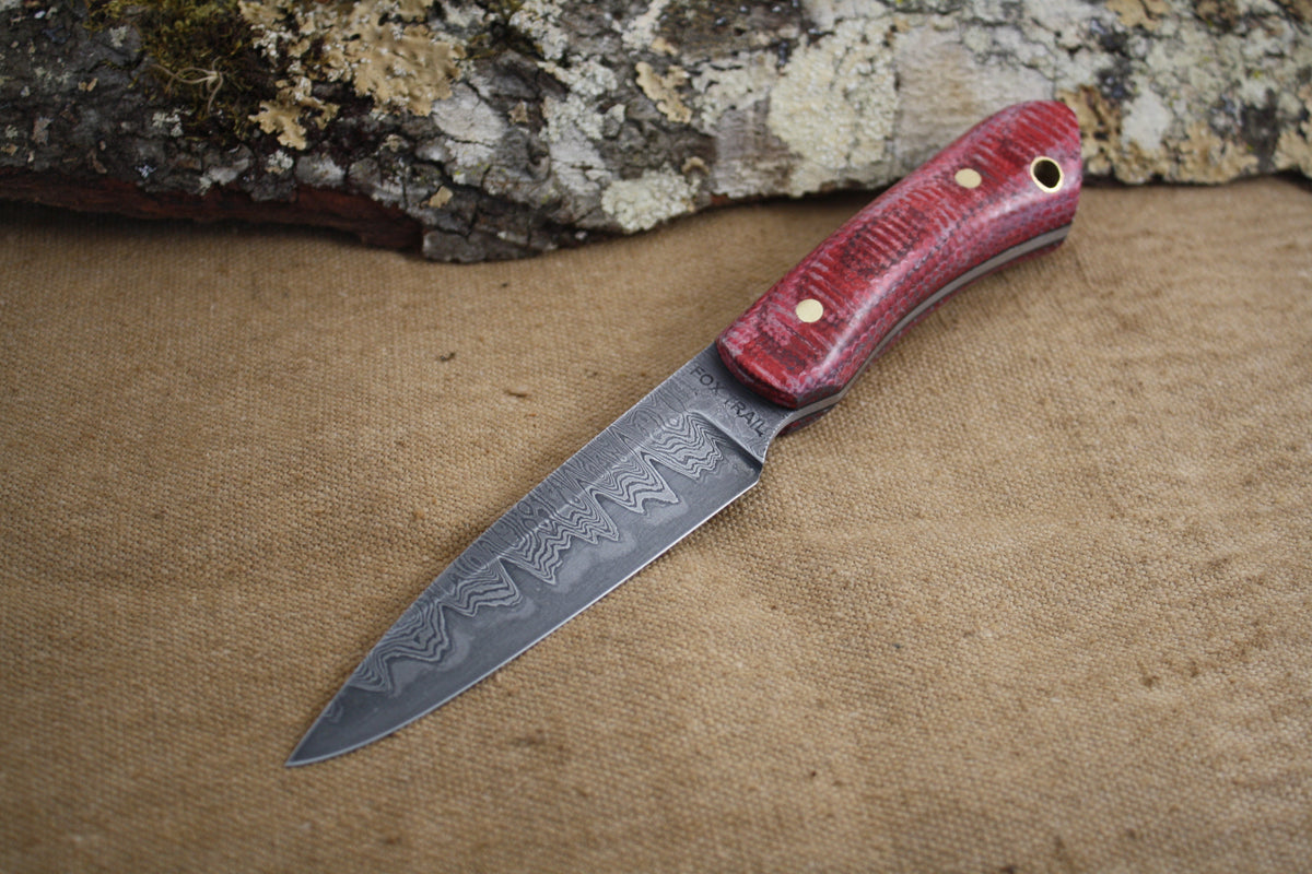 Hand‑forged raindrop Damascus knife made from fire sprinkler pipe and 1095 steel with micarta fire hose handle, created for the Buddy Dewar Golf and Skeet Tournament raffle, displayed on burlap background with natural bark accent.