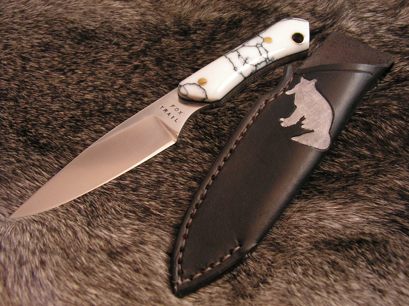 Neck knife displayed on fur, featuring a Snow Fox 3 stainless steel blade with a white synthetic marble handle veined in dark gray. Shown with leather sheath, detailed with a fox-shaped cutout.