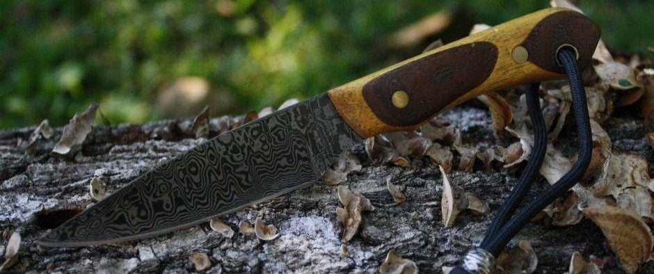 Hand‑forged Damascus neck knife with random pattern blade, stacked Yellowheart and walnut handle, leather thong, and brass pins, displayed on a log with grass in the background.