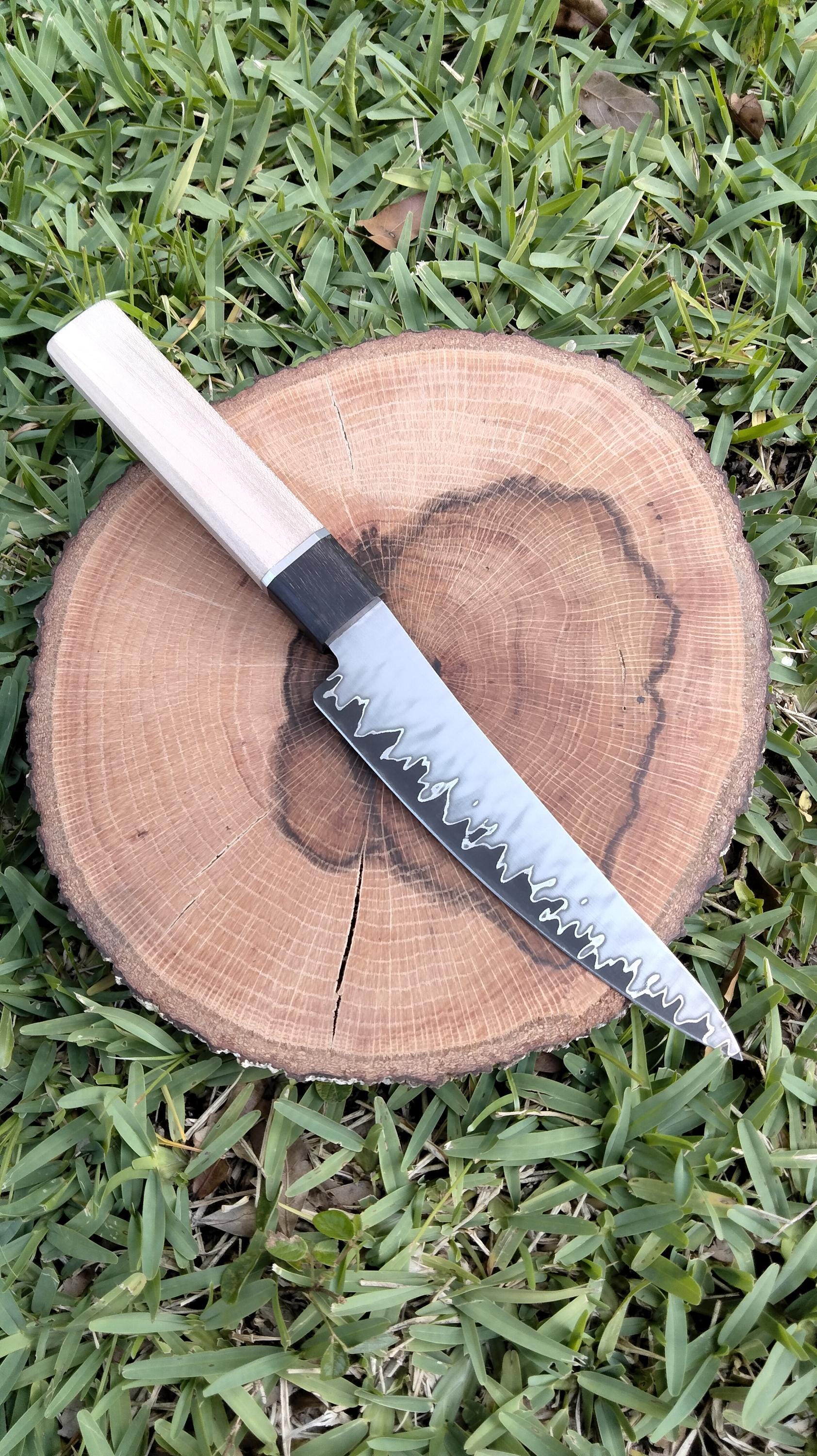 Top view of opposite side of San‑Mai utility knife on wood with grass background, highlighting striking layered pattern, curved spine, and hand‑crafted handle details.