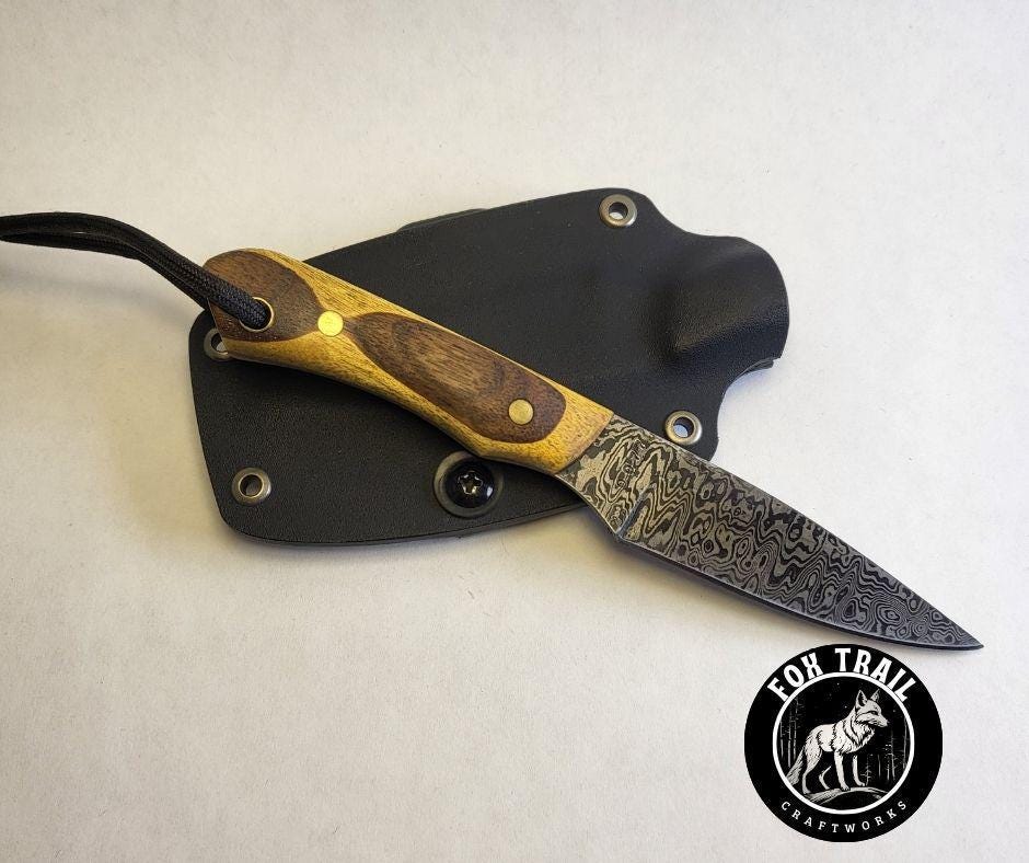 Hand‑forged Damascus neck knife with stacked Yellowheart and walnut handle, brass pins, and black wrist thong, leaning against black Kydex sheath on white background.