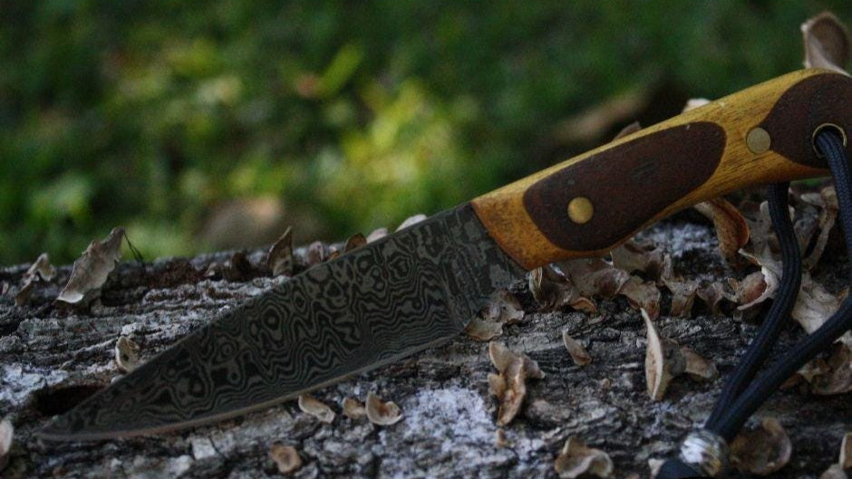 Hand‑forged Damascus neck knife resting on log with grass backdrop, handle right side, makers mark visible on blade, showing Yellowheart and walnut handle and black wrist thong.