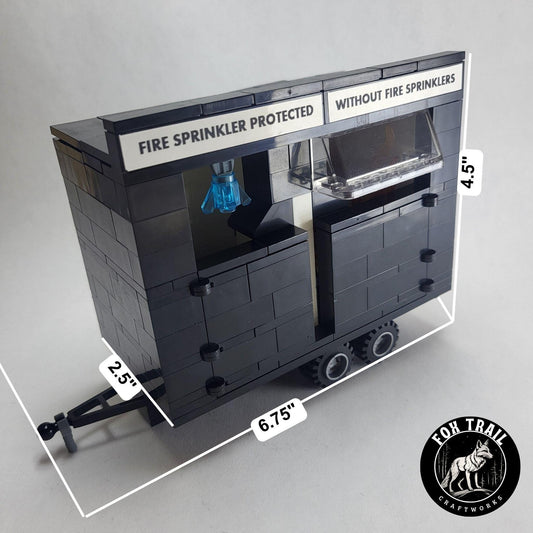 Left‑side view of Lego‑compatible burn trailer with doors closed, trailer coupler, and signage comparing fire sprinkler protection versus no sprinklers. The windscreen on the unprotected side mirrors real burn trailer designs used in fire safety demos.