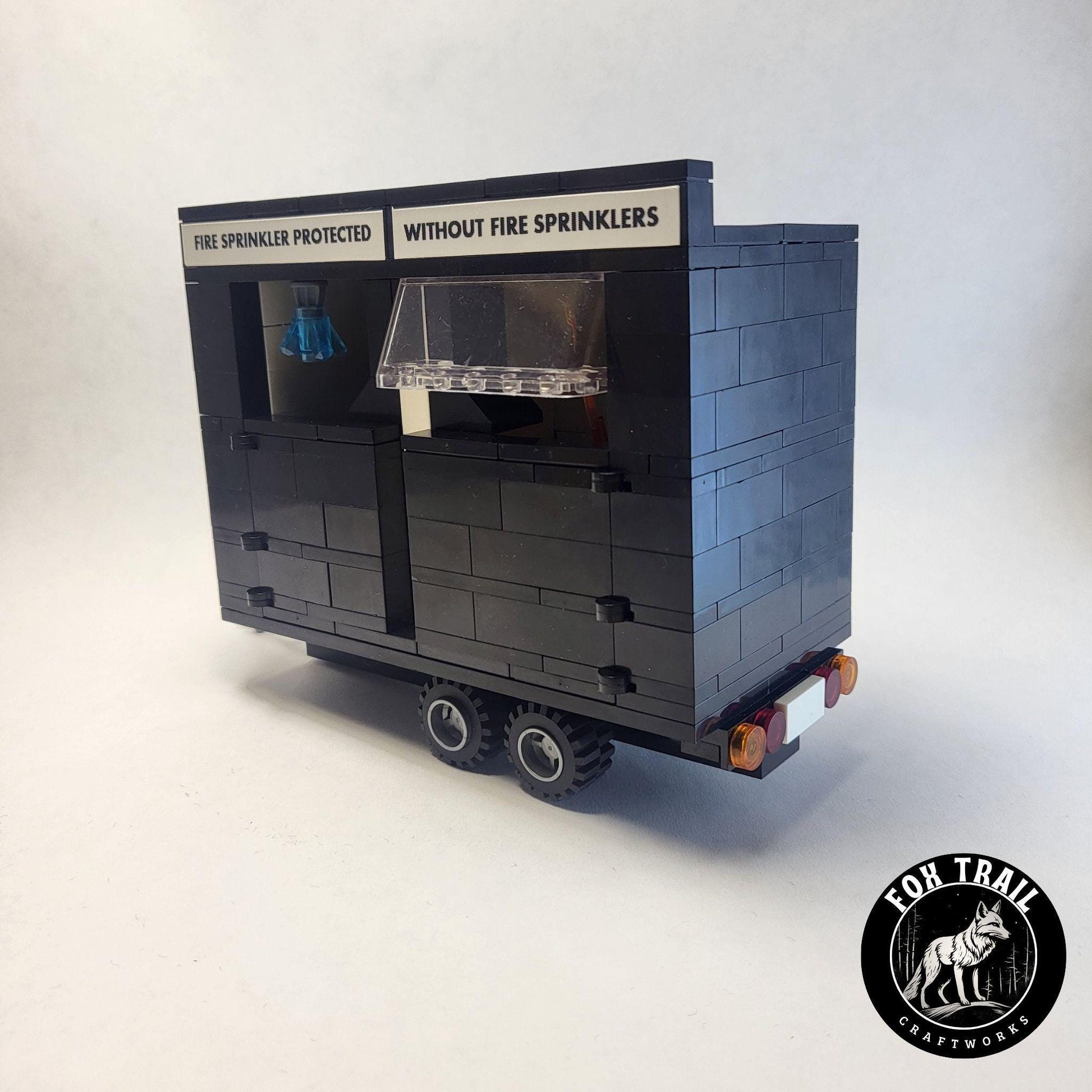 Right‑side view of Lego‑style fire sprinkler burn trailer showing tail lights, license plate detail, and signage contrasting ‘Fire Sprinkler Protected’ and ‘Without Fire Sprinklers’ sides for educational demonstrations.