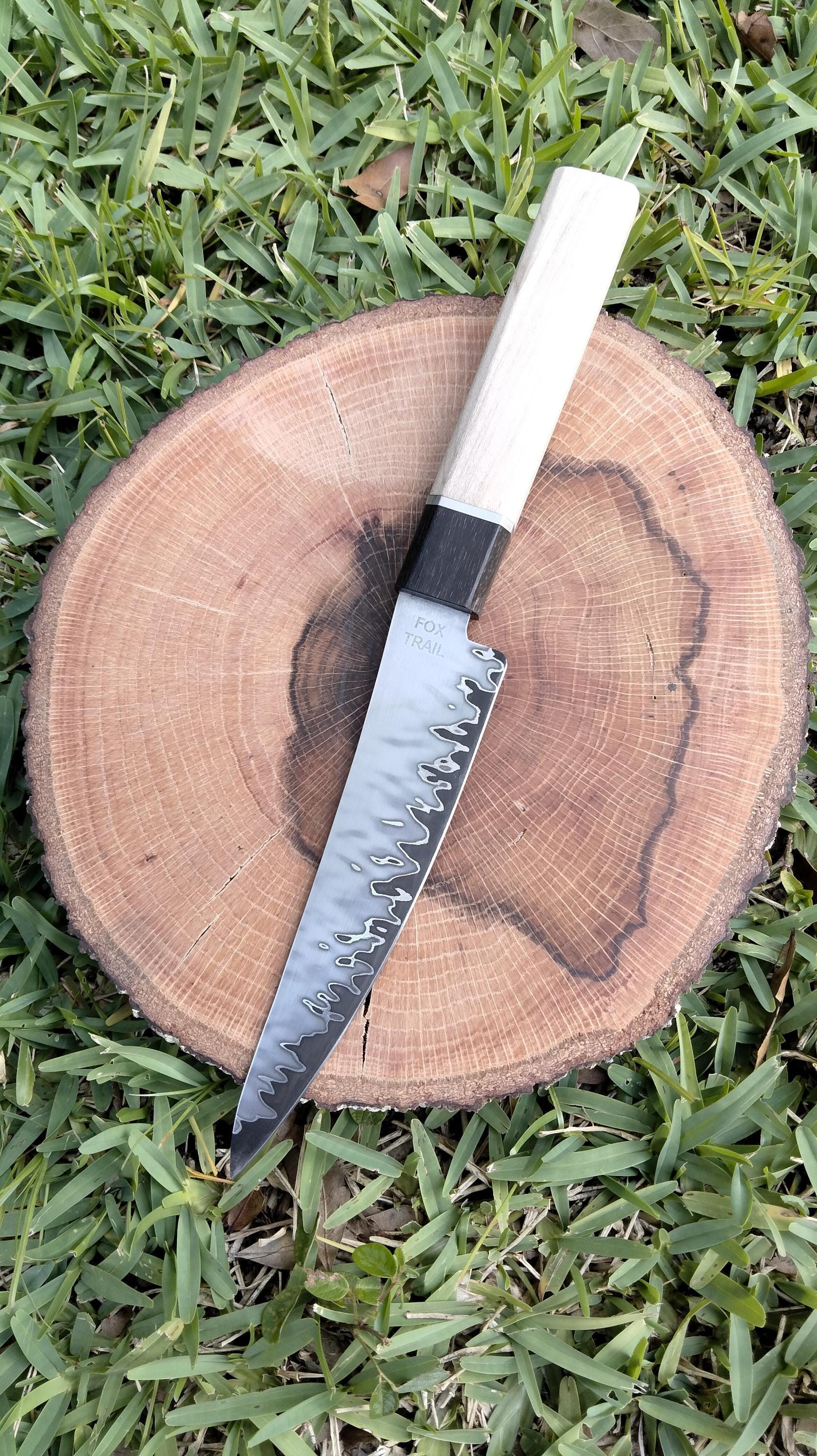 Top view of hand‑forged San‑Mai utility knife on wood with grass background, featuring Fox Trail Craftworks maker’s mark and curved spine; striking Damascus‑style pattern with Gaboon ebony and silver maple handle visible.