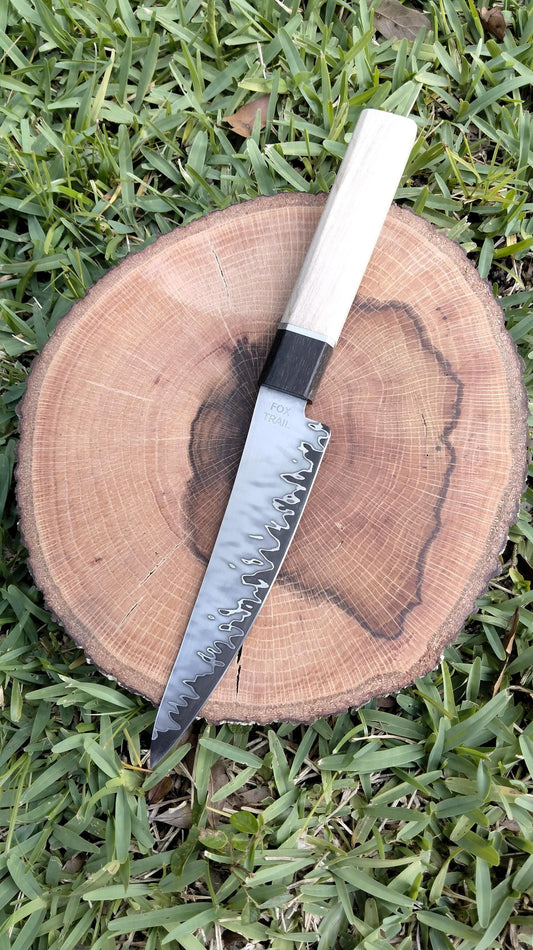 Top view of hand‑forged San‑Mai utility knife on wood with grass background, featuring Fox Trail Craftworks maker’s mark and curved spine; striking Damascus‑style pattern with Gaboon ebony and silver maple handle visible.