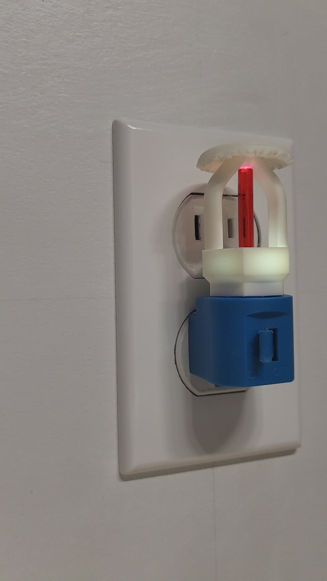 Video of fire sprinkler night light plugged into white wall outlet with switch on, glowing softly in a well‑lit room as the view rotates 180 degrees to show left, front, and right angles of the white upright head with red bulb on blue base.
