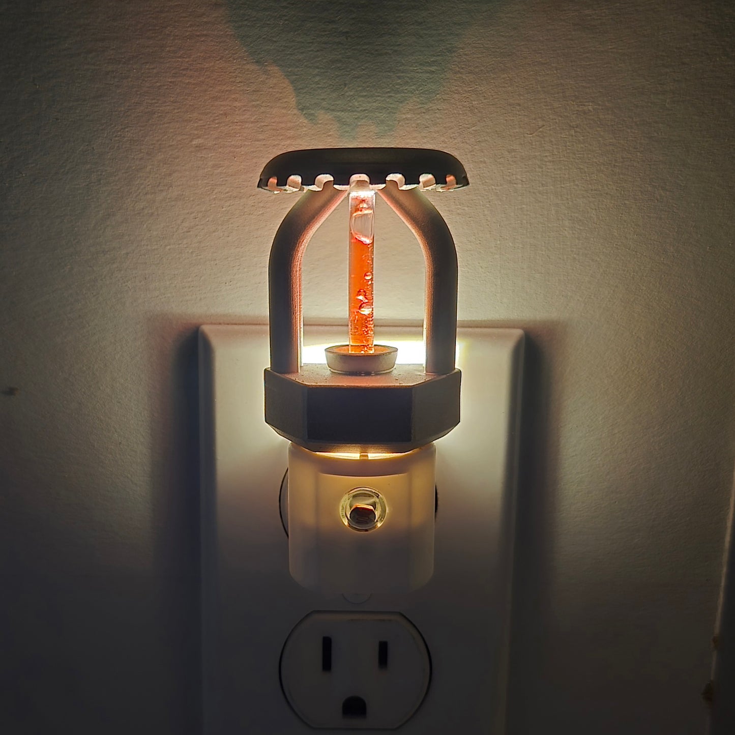Front close‑up of realistic fire sprinkler night light plugged in and glowing, nickel‑plated head casting a soft shadow on the wall, red bulb detail reflecting residential fire protection style