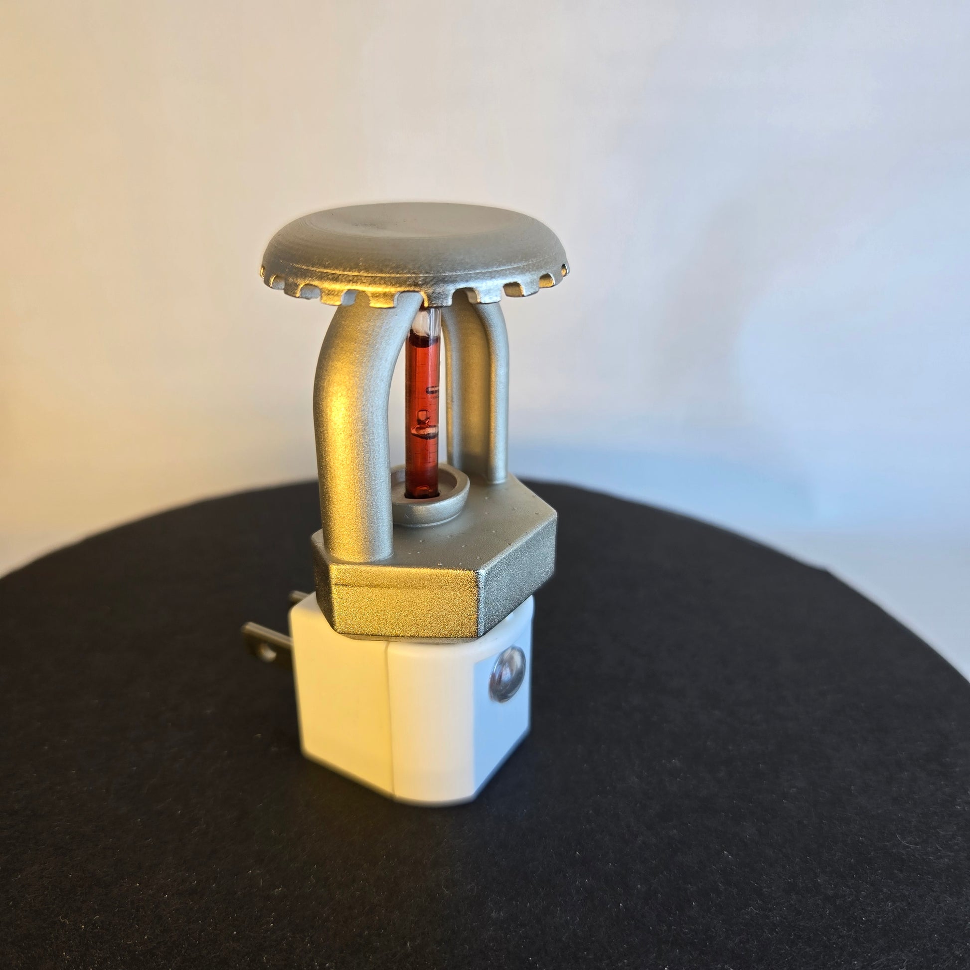 Left‑side view of upright fire sprinkler night light with metallic nickel finish and red bulb, mounted on white night light base with dusk‑to‑dawn sensor, displayed on black stand against white backdrop.