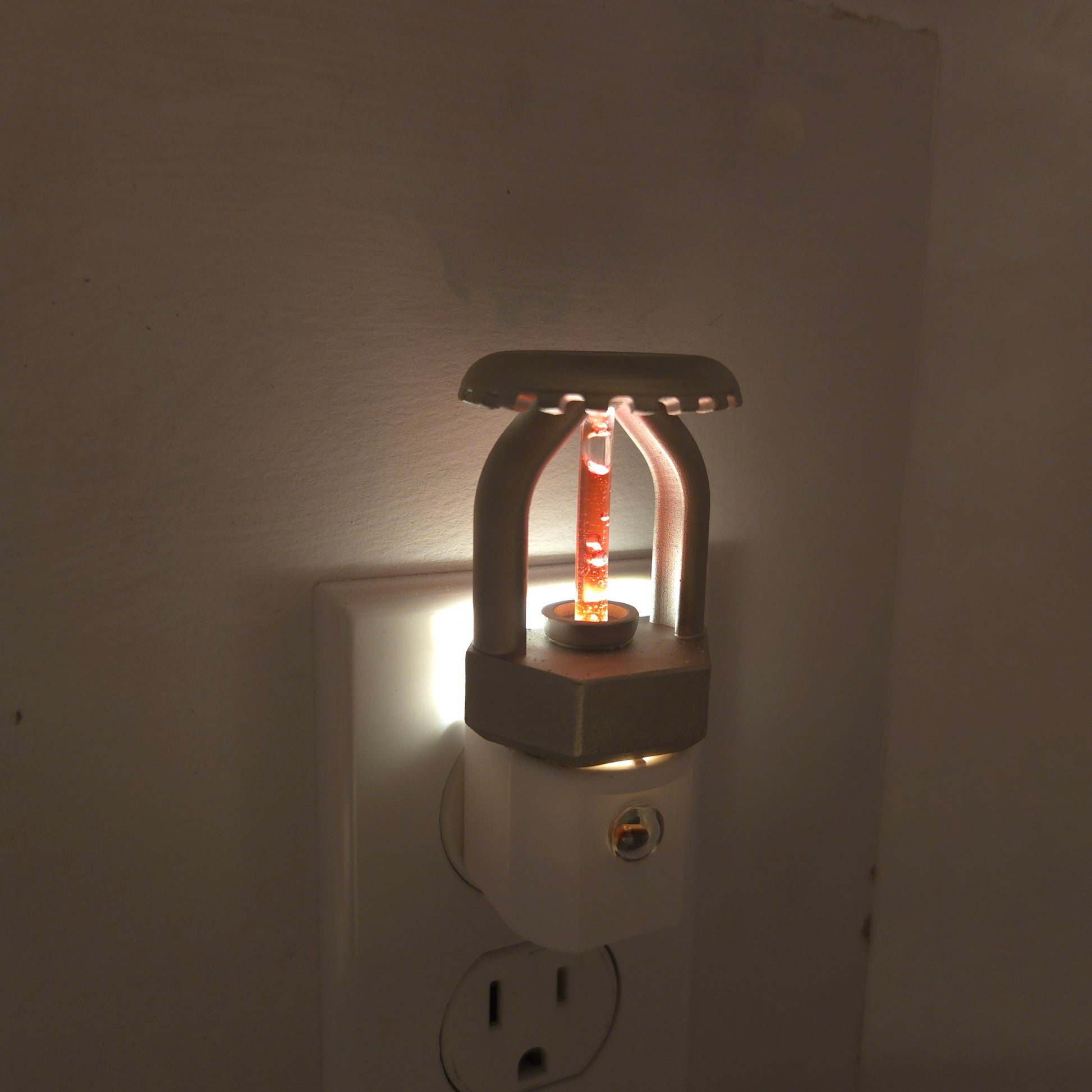 Realistic fire sprinkler night light plugged into outlet in dark room, LED softly glowing to show red residential bulb and nickel‑plated head; dusk‑to‑dawn sensor automatically activates in low light.