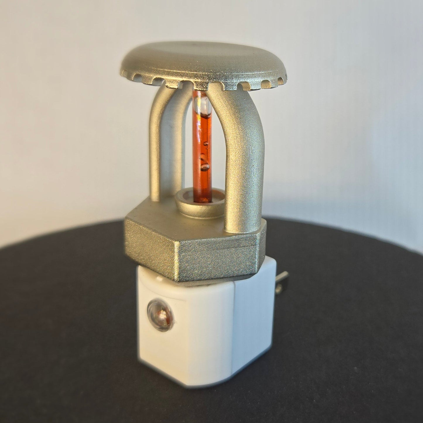 Right‑side view of realistic fire sprinkler night light on white background, showing nickel‑plated upright sprinkler head with red liquid bulb, white night light base with dusk‑to‑dawn sensor, and black display disc.