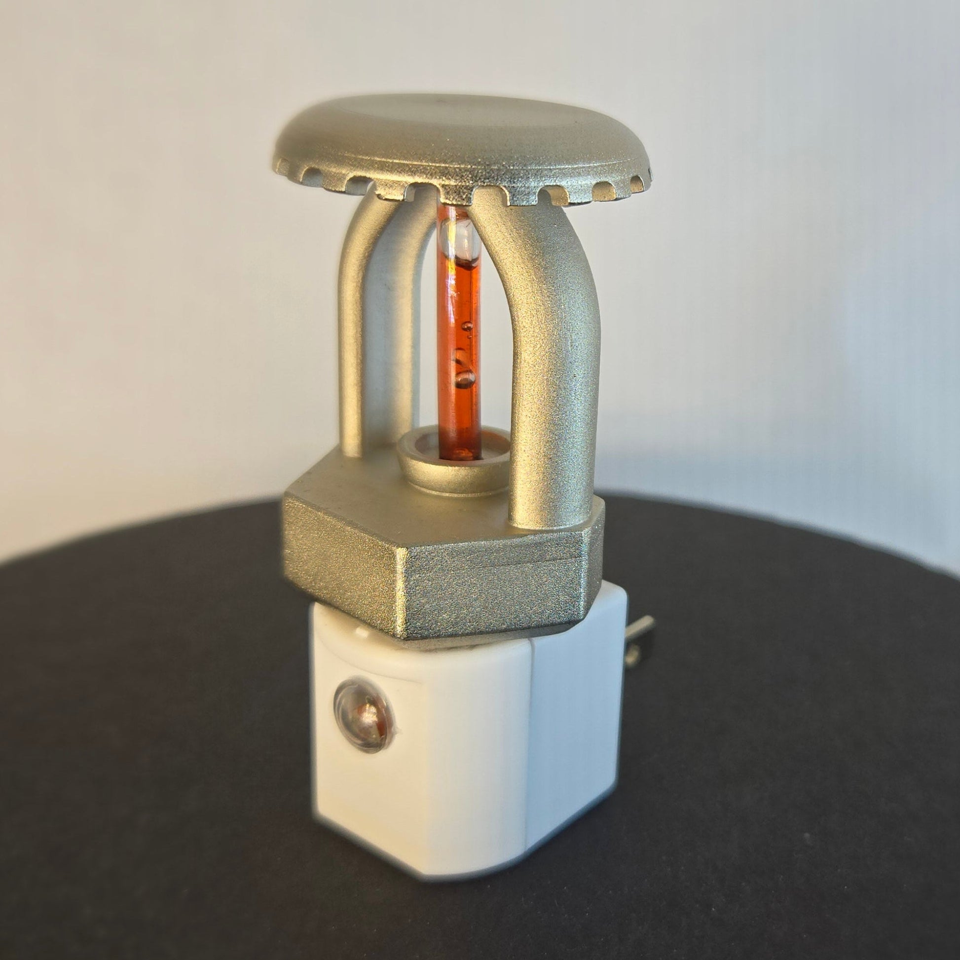 Right‑side view of realistic fire sprinkler night light on white background, showing nickel‑plated upright sprinkler head with red liquid bulb, white night light base with dusk‑to‑dawn sensor, and black display disc.