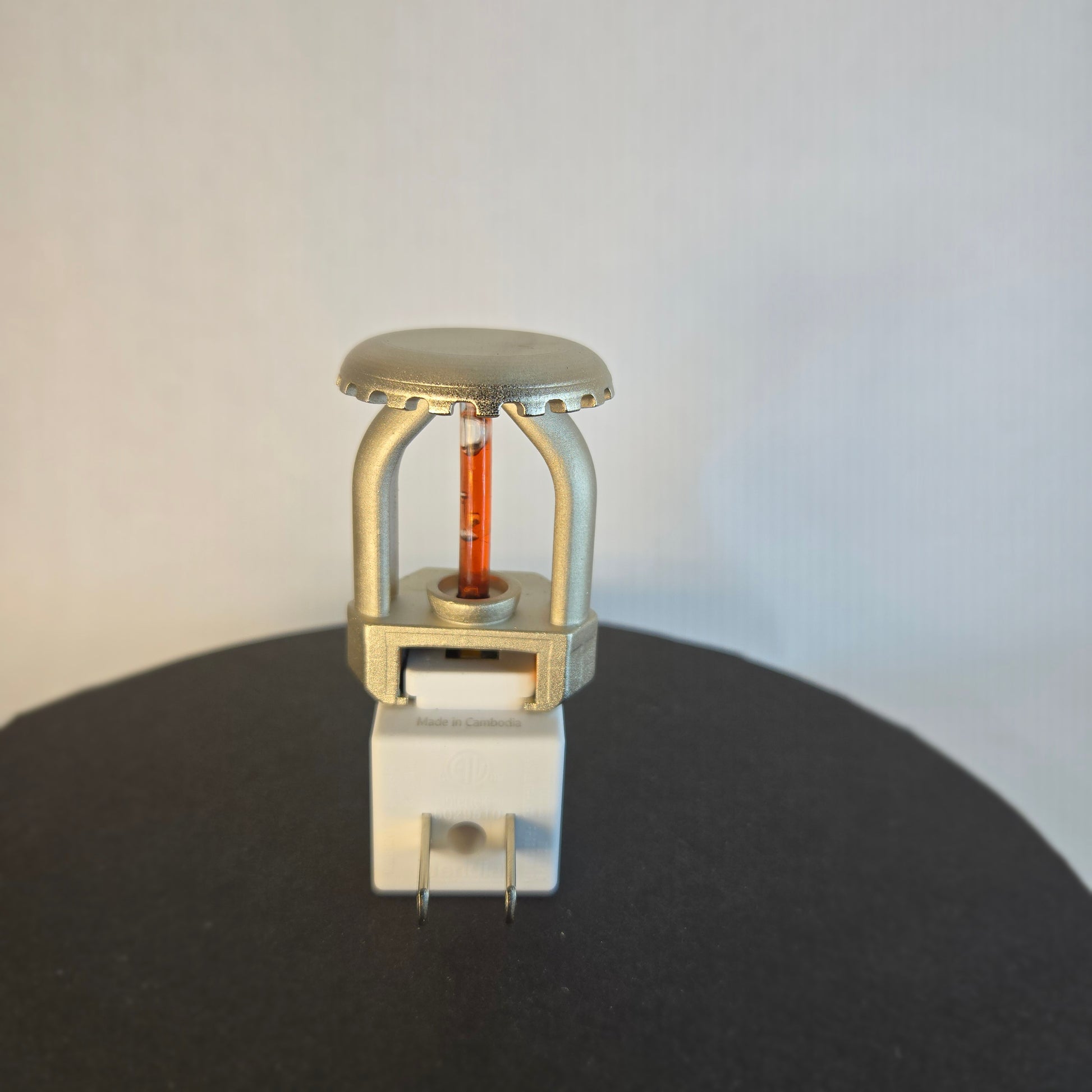 Back view of realistic fire sprinkler night light with nickel‑plated upright head and red bulb, white night light housing, and standard two‑prong plug for household outlets.