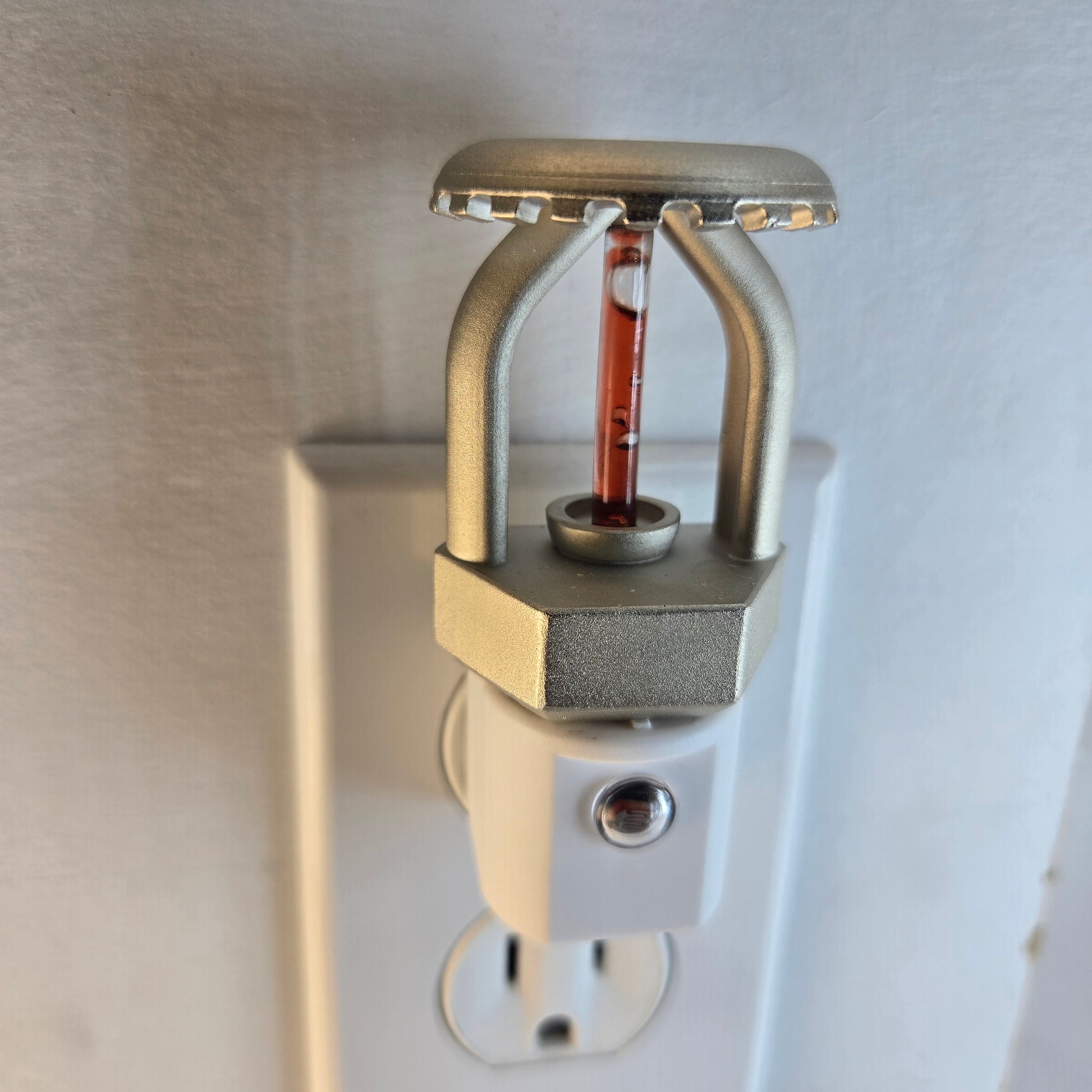 Left‑side view of upright fire sprinkler night light plugged into outlet on white wall in daylight; LED is off, showing nickel‑plated head, red bulb, and built‑in dusk‑to‑dawn sensor.