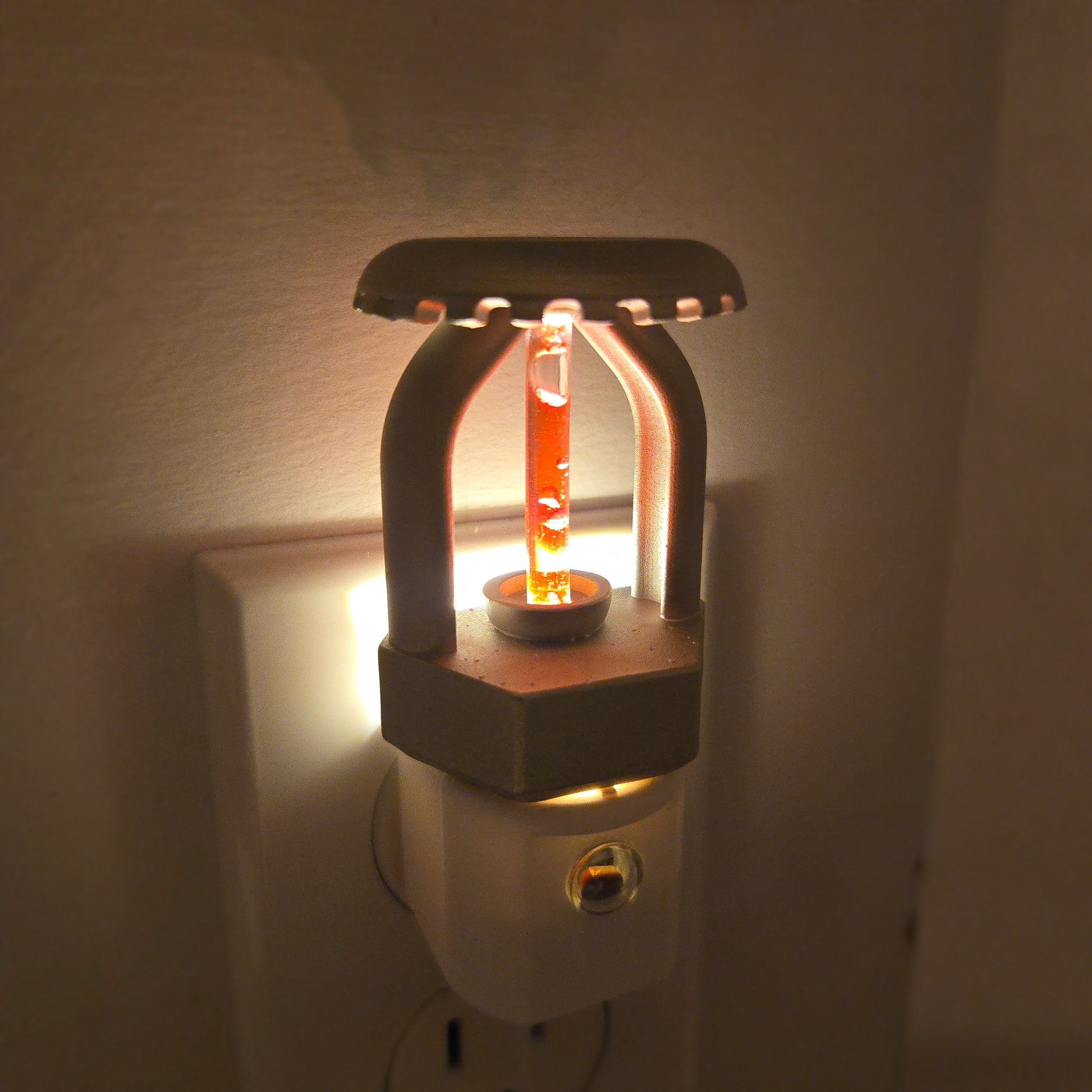 Close‑up of realistic fire sprinkler night light plugged into outlet at night, highlighting nickel‑plated finish, red bulb detail, and warm glow that adds subtle fire‑protection charm to any room.