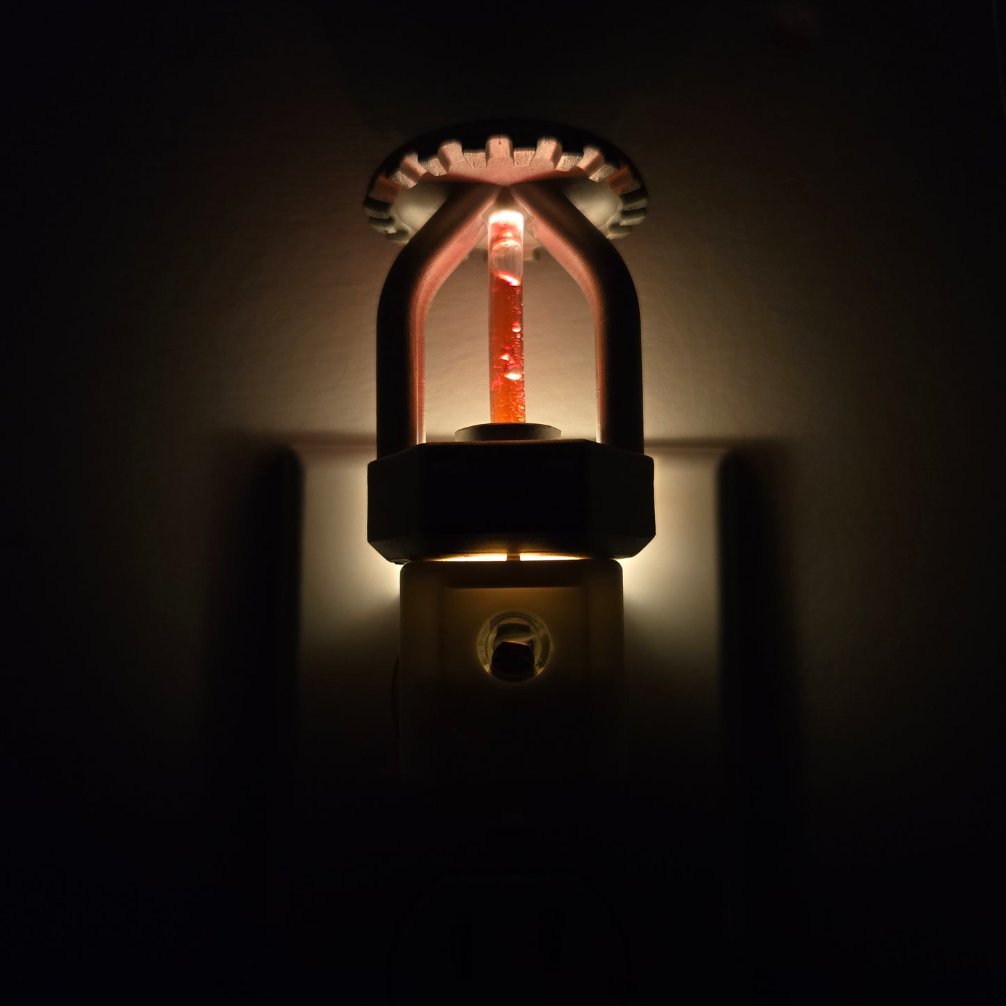 Front close‑up of realistic fire sprinkler night light plugged in and glowing in dark space, showing red liquid‑style bulb and nickel finish that mirrors real residential sprinklers.