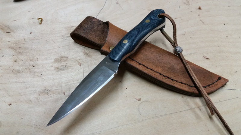 Neck knife with 440C stainless steel blade and micarta‑style denim handle, paired with a pouch‑style leather sheath and leather wrist thong with bead, displayed on plywood.