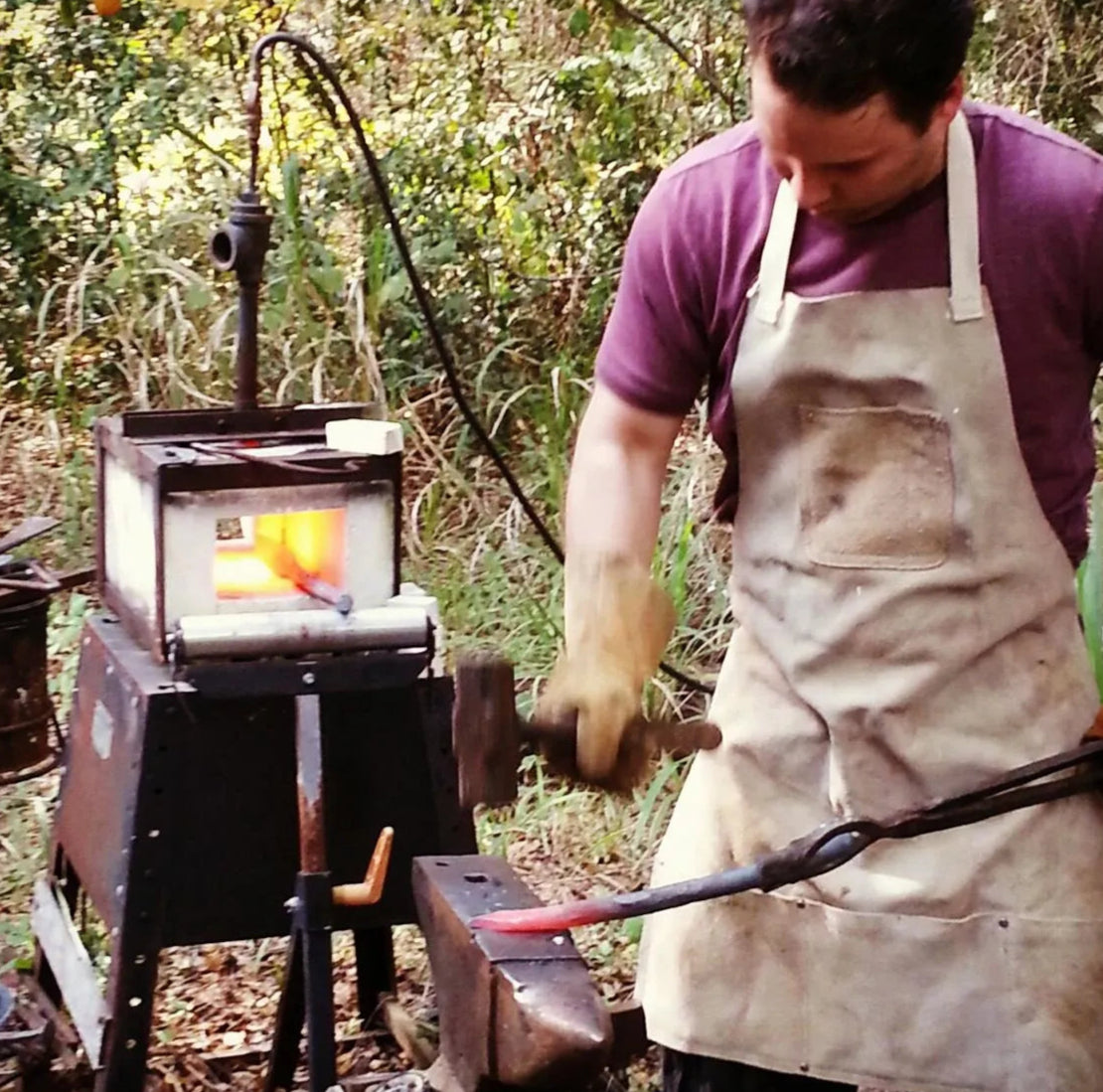 Chris forging a knife on a small anvil in his backyard, wearing a leather apron. An early forge setup is visible in the background in this older photo.

