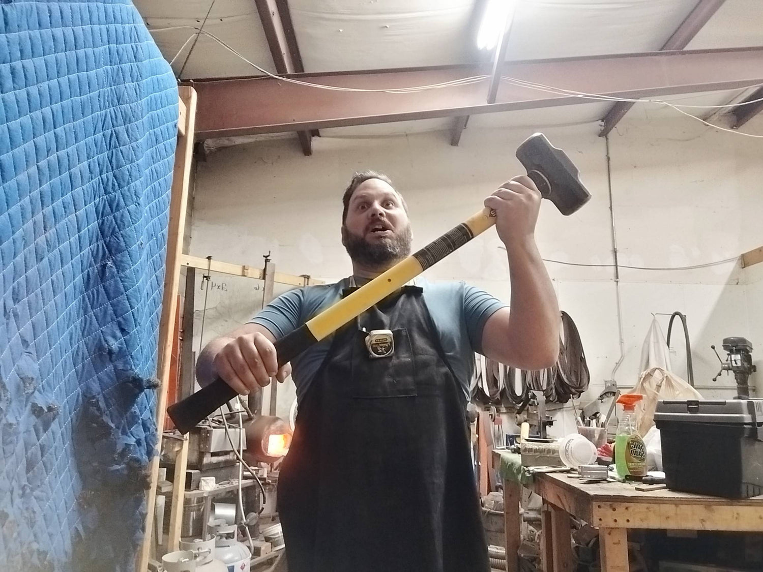 Chris in the Fox Trail Craftworks workshop wearing a leather apron, holding a sledgehammer in a playful ‘Thor’ pose while bladesmithing.