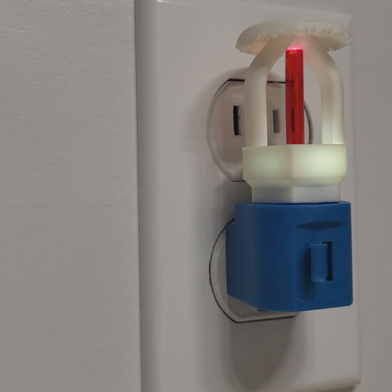 Video of fire sprinkler night light plugged into white wall outlet with switch on, glowing softly in a well‑lit room as the view rotates 180 degrees to show left, front, and right angles of the white upright head with red bulb on blue base.