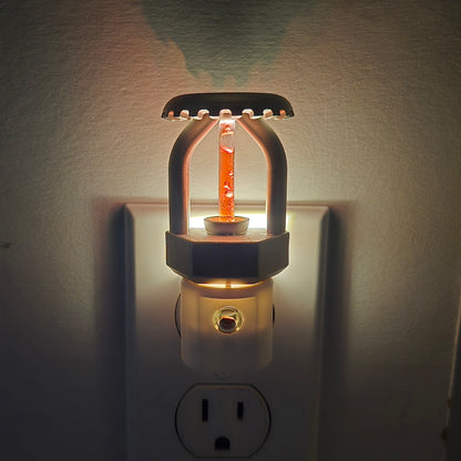 Front close‑up of realistic fire sprinkler night light plugged in and glowing, nickel‑plated head casting a soft shadow on the wall, red bulb detail reflecting residential fire protection style