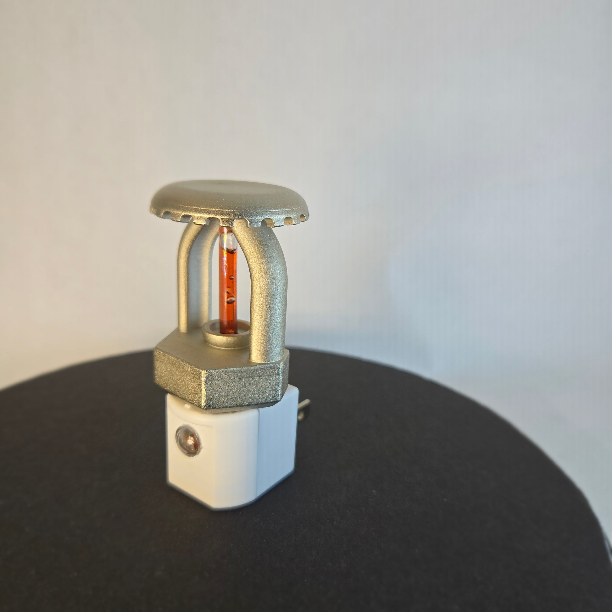 Right‑side view of realistic fire sprinkler night light on white background, showing nickel‑plated upright sprinkler head with red liquid bulb, white night light base with dusk‑to‑dawn sensor, and black display disc.