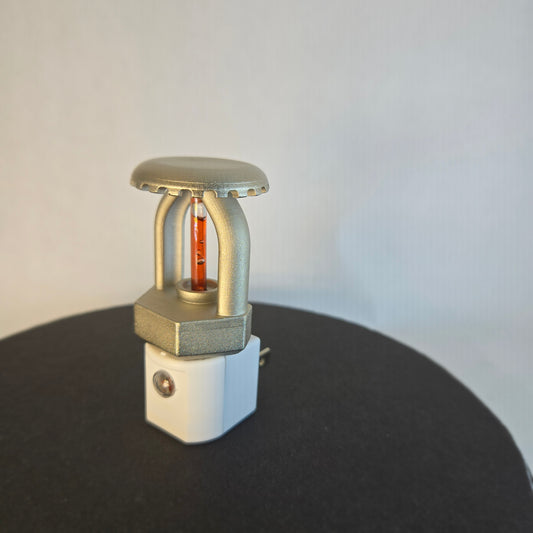 Right‑side view of realistic fire sprinkler night light on white background, showing nickel‑plated upright sprinkler head with red liquid bulb, white night light base with dusk‑to‑dawn sensor, and black display disc.
